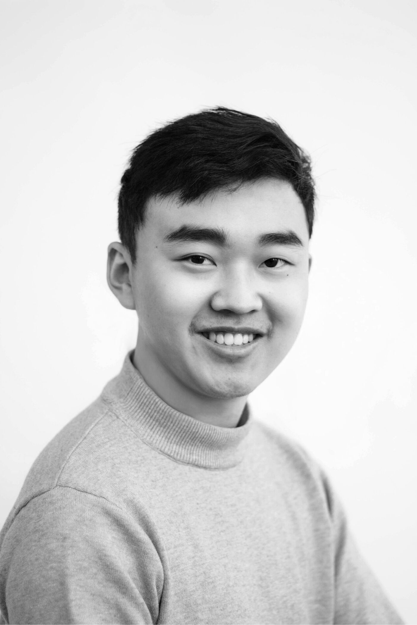 Close-up black and white shot of a young man with short dark hair, wearing a grey mock turtleneck sweater, smiling gently at the camera.