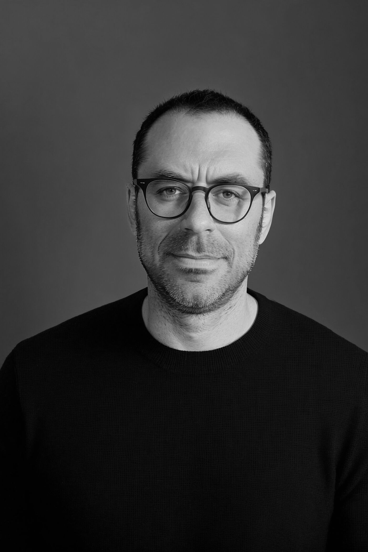 Close-up black and white portrait of a man with glasses wearing a black crewneck sweater against a gray background.