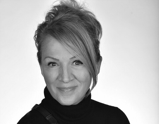 Close-up black and white shot of a woman with short, slightly tousled hair, wearing a black turtleneck and a crossbody bag, her hands clasped in front of her.
