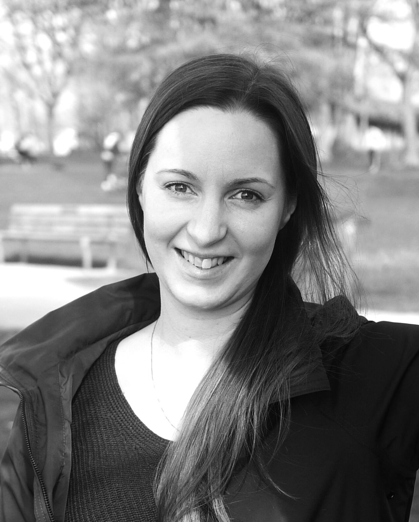 A black and white, eye-level, medium shot of a woman with long brown hair, wearing a dark jacket and smiling.