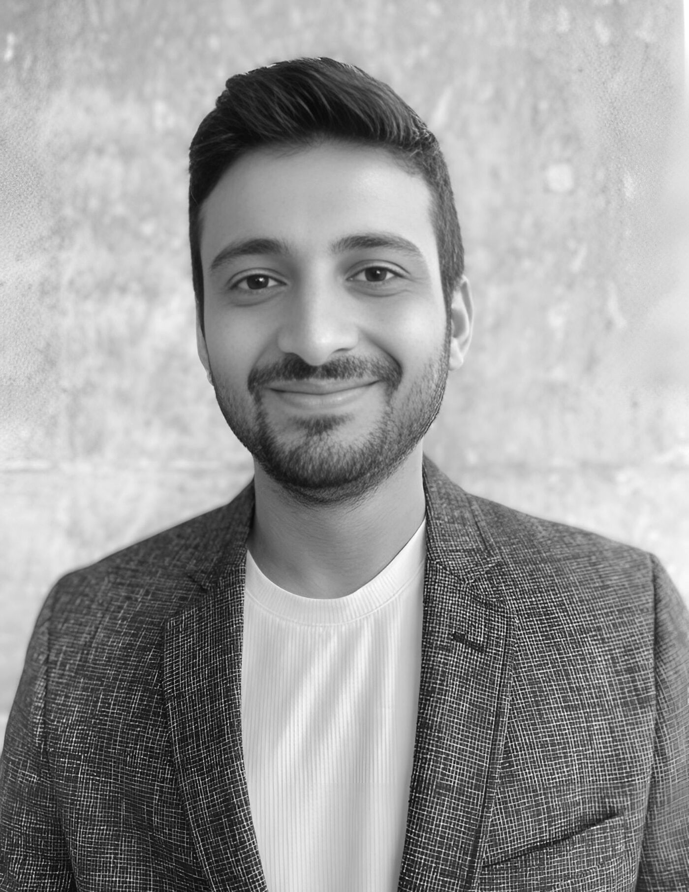Close-up black and white shot of a man with short brown hair, wearing a textured blazer and a white t-shirt, smiling subtly against a mottled background.
