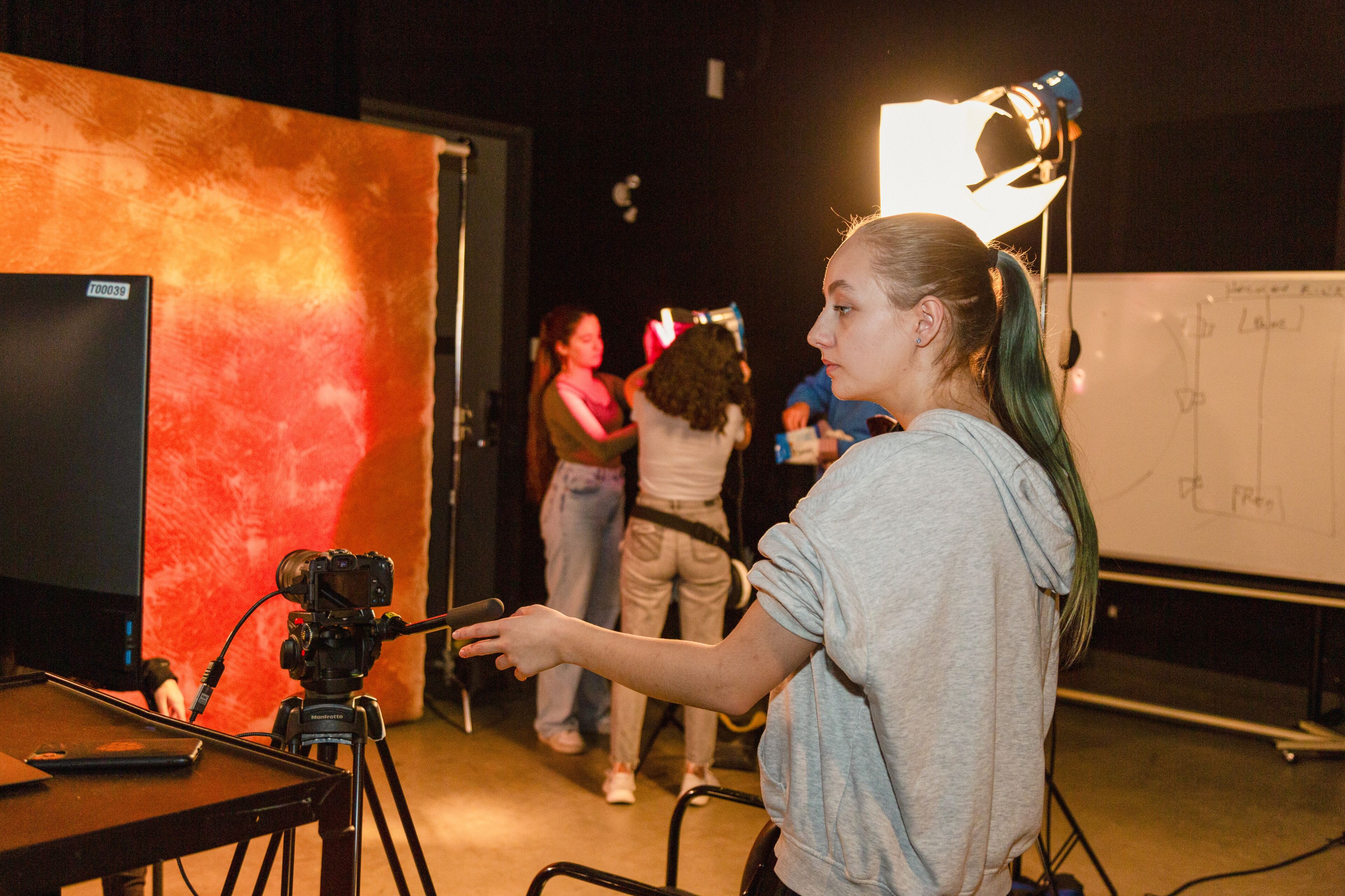 A woman directs a scene, pointing towards a camera on a tripod with studio lights and participants in the background.