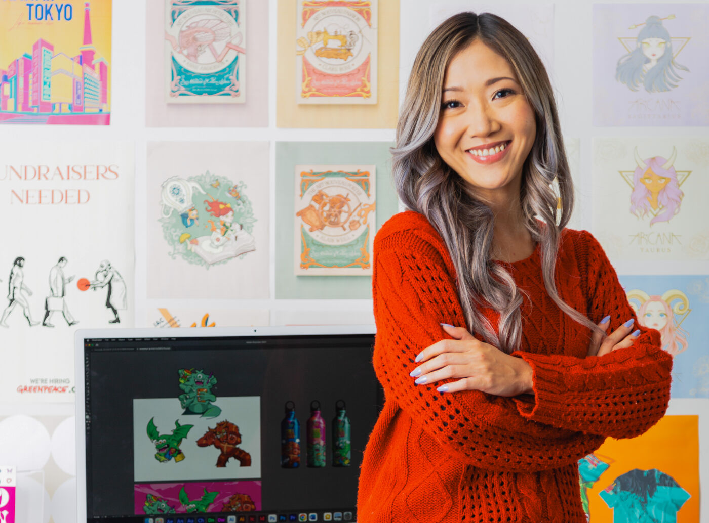 A smiling woman in a red sweater stands before her colorful artwork, which includes character illustrations and graphics on the wall and computer.