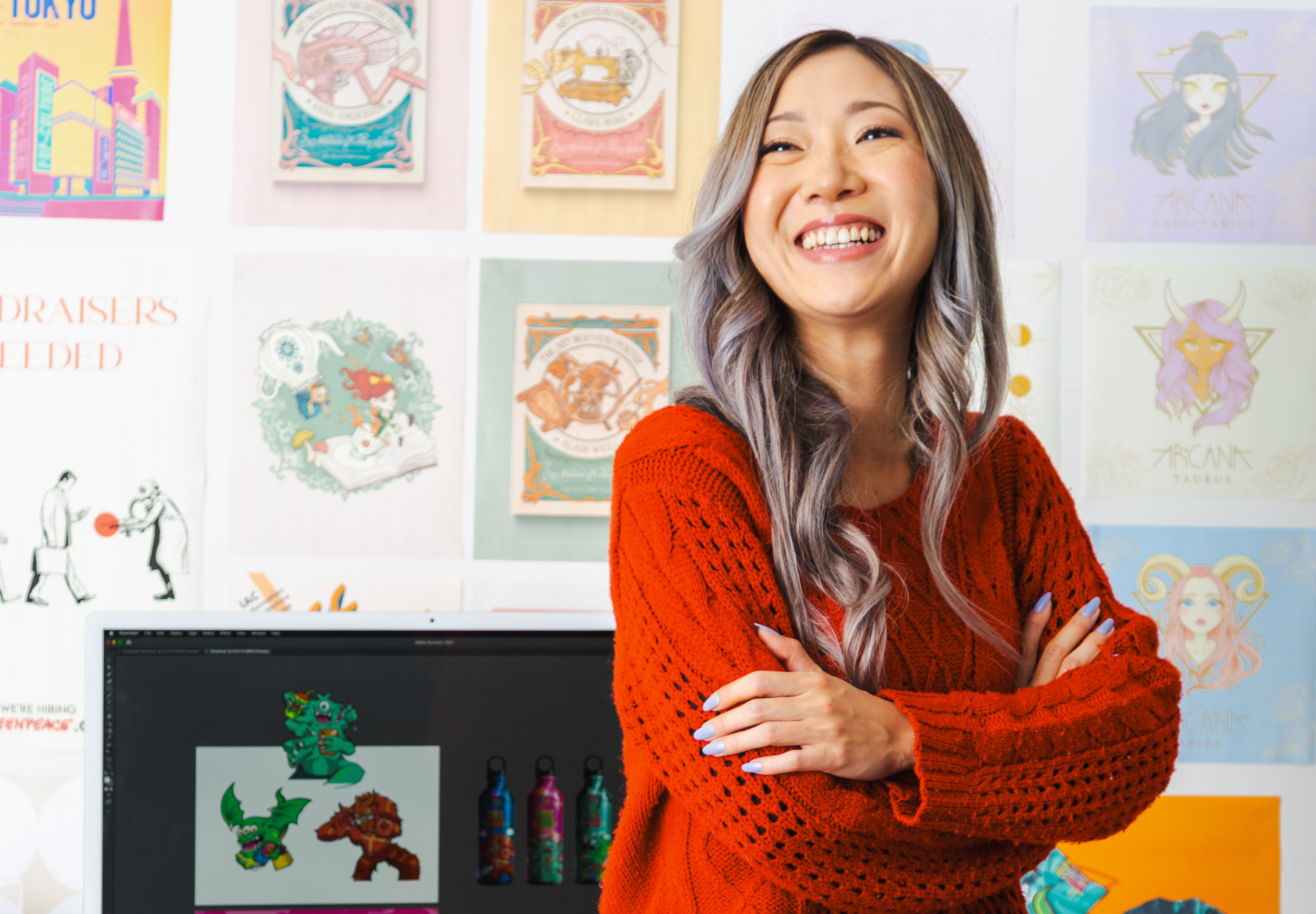 Une artiste joyeuse aux cheveux argentés en pull rouge se tient dans son atelier entourée d'illustrations colorées.