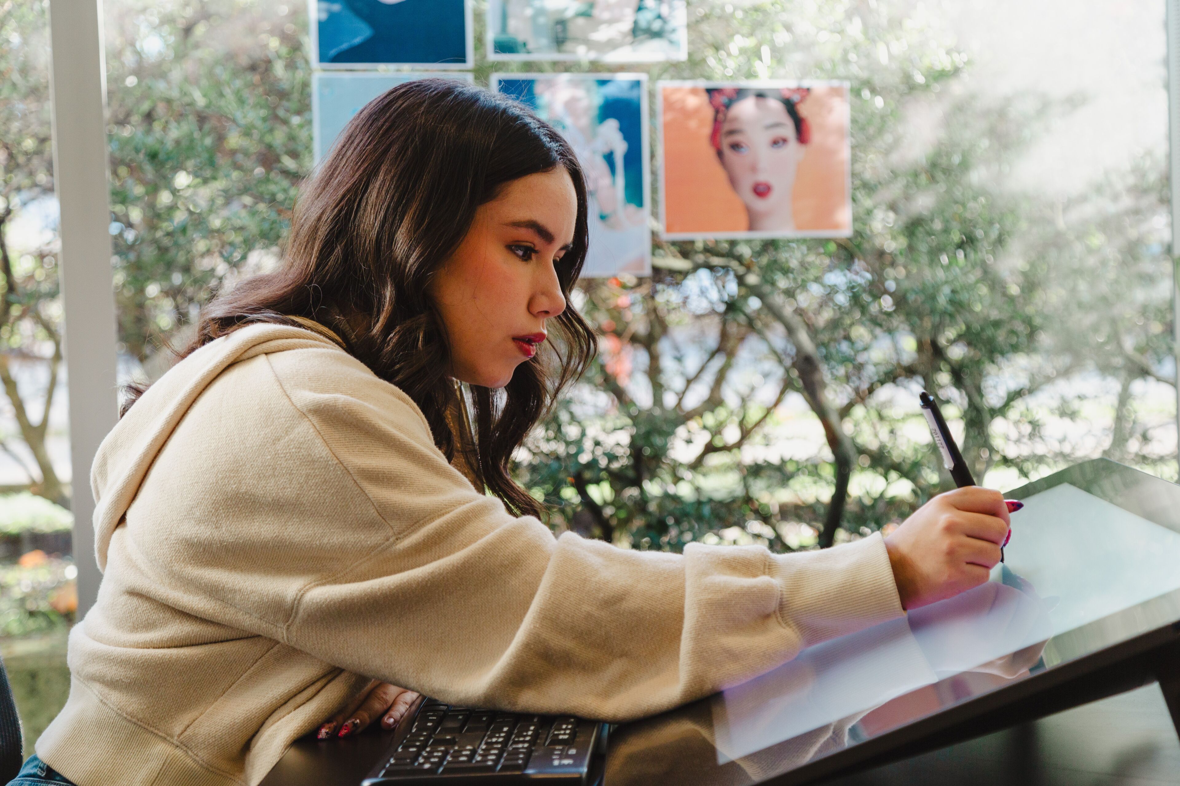 A focused artist sketches on a digital tablet by a window with inspiration images.