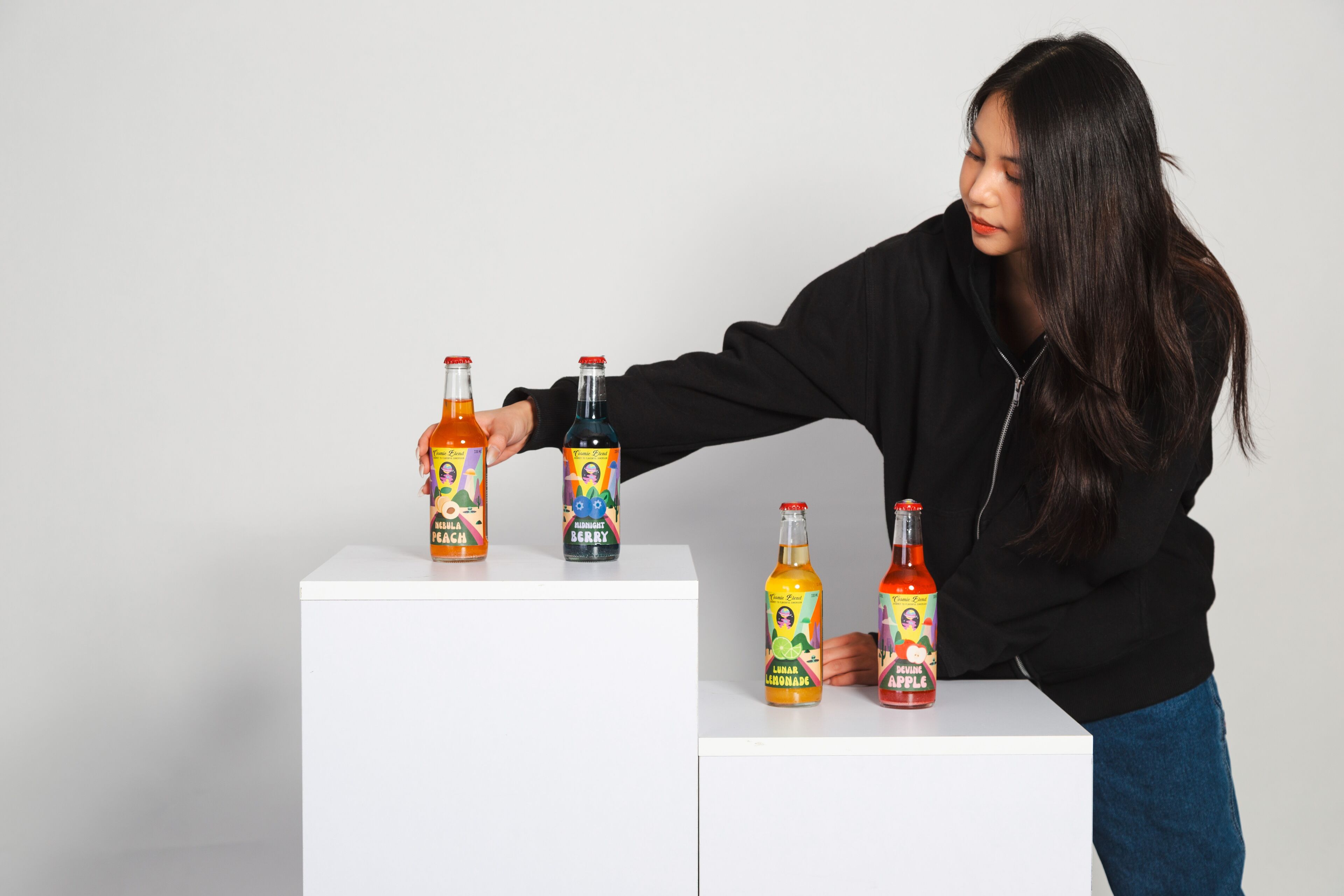 A young woman in casual attire arranges bottles with vibrant label designs, demonstrating her graphic design skills on a white display.
