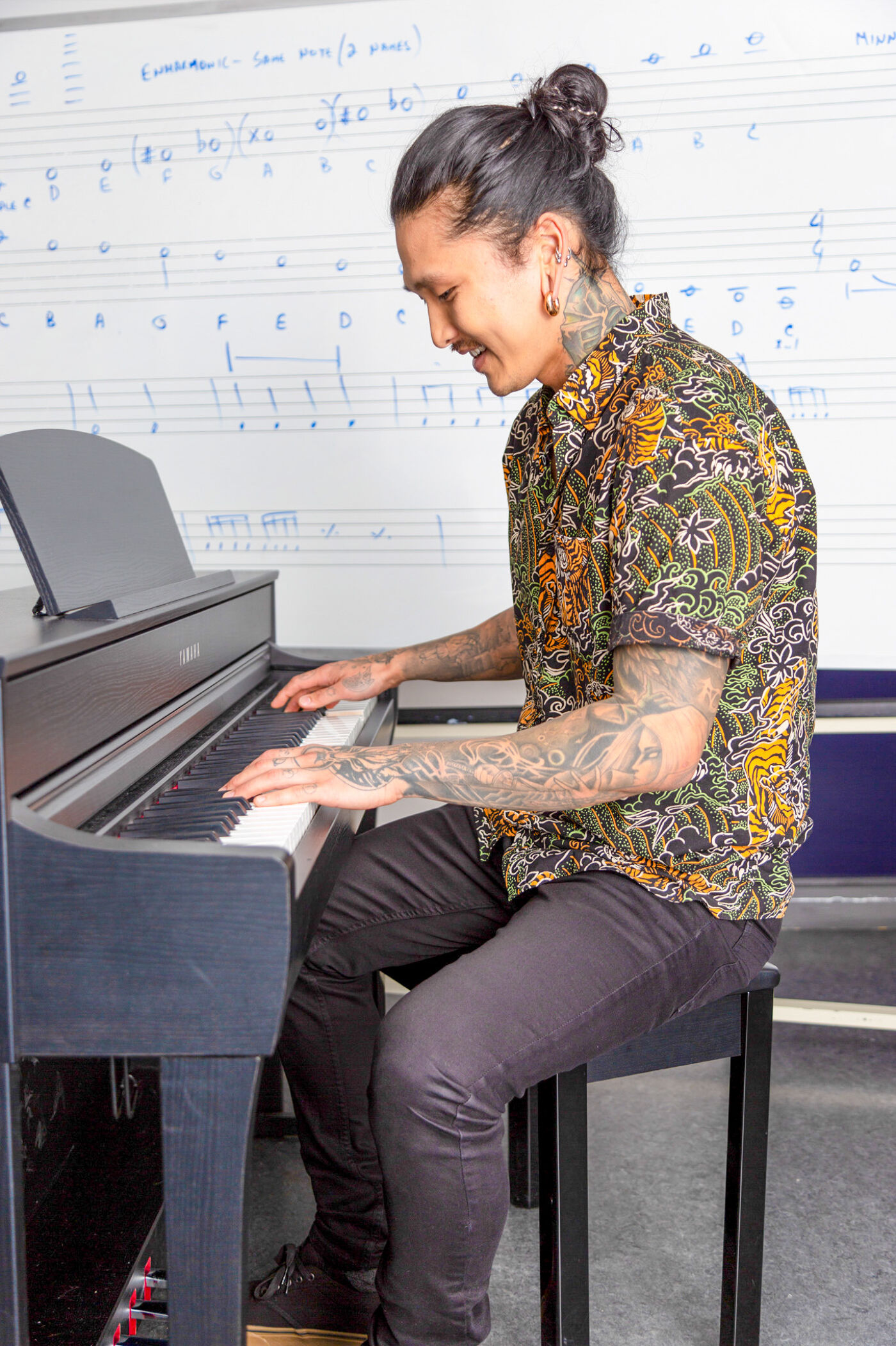 A person with sleeve tattoos and a topknot hairstyle is playing a digital piano, with musical scores in the background.