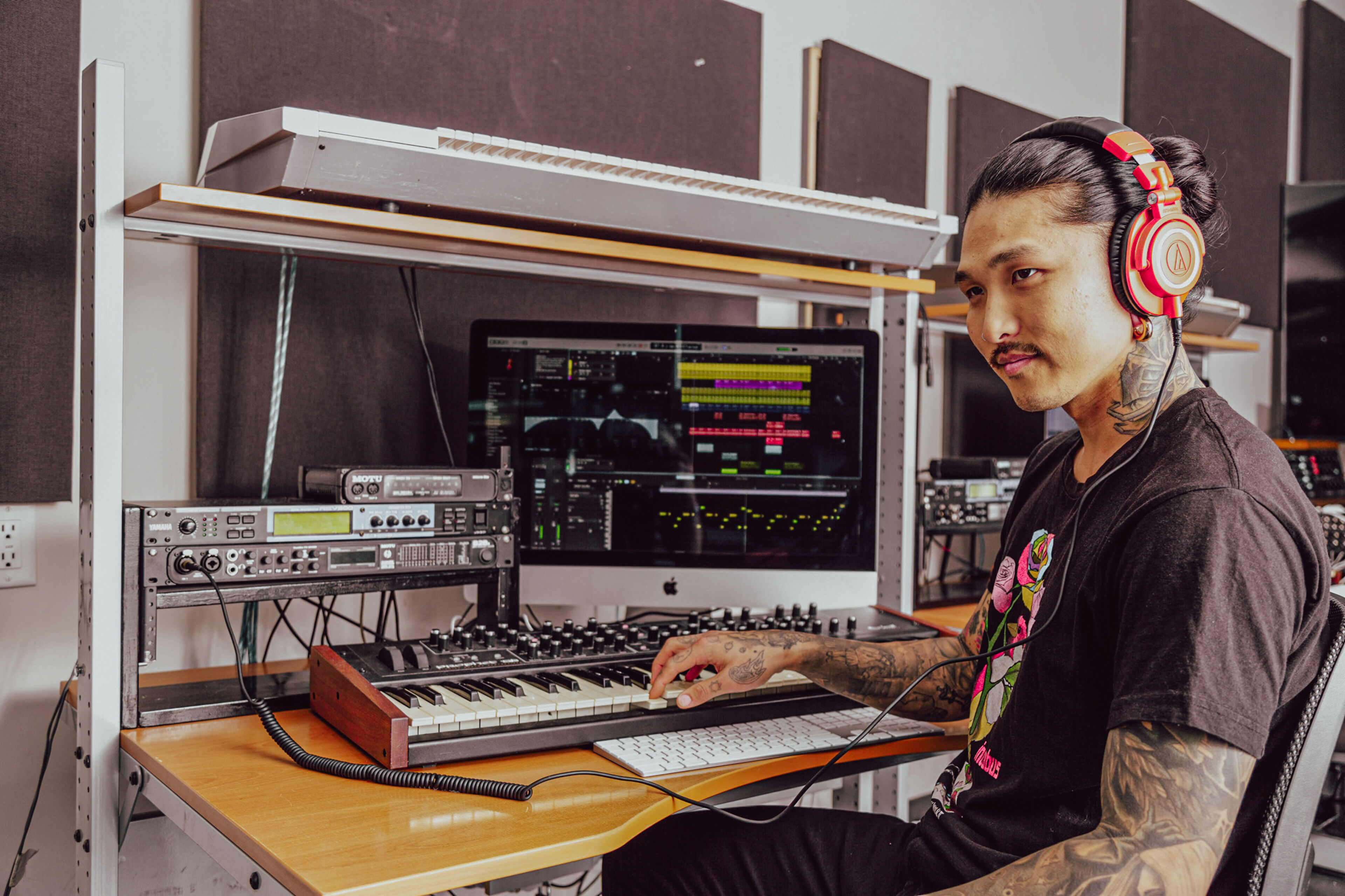 A tattooed music producer is focused on mixing tracks in a studio, with a MIDI keyboard and audio equipment surrounding him.
