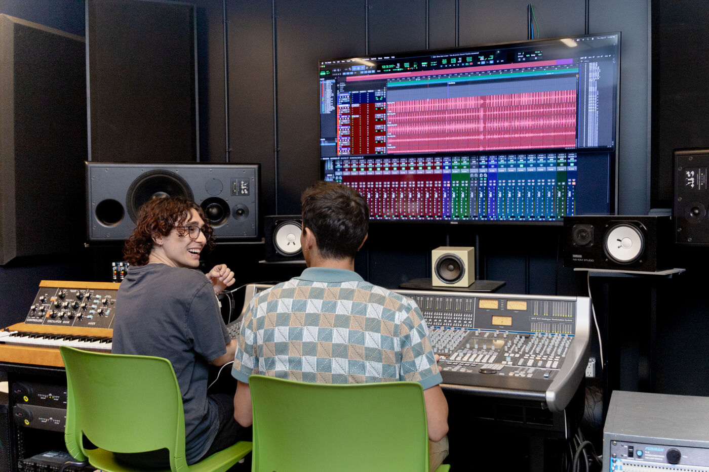 Two people are working in a recording studio, with a large mixing board and a screen displaying music production software.