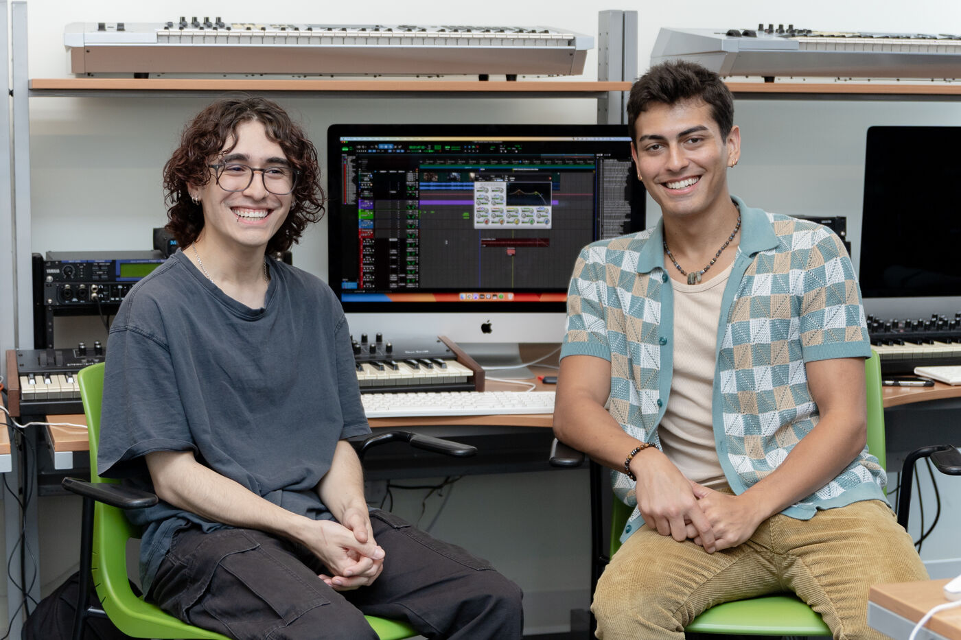 Two smiling young men are seated in front of a computer in a music studio, surrounded by keyboards and recording equipment.