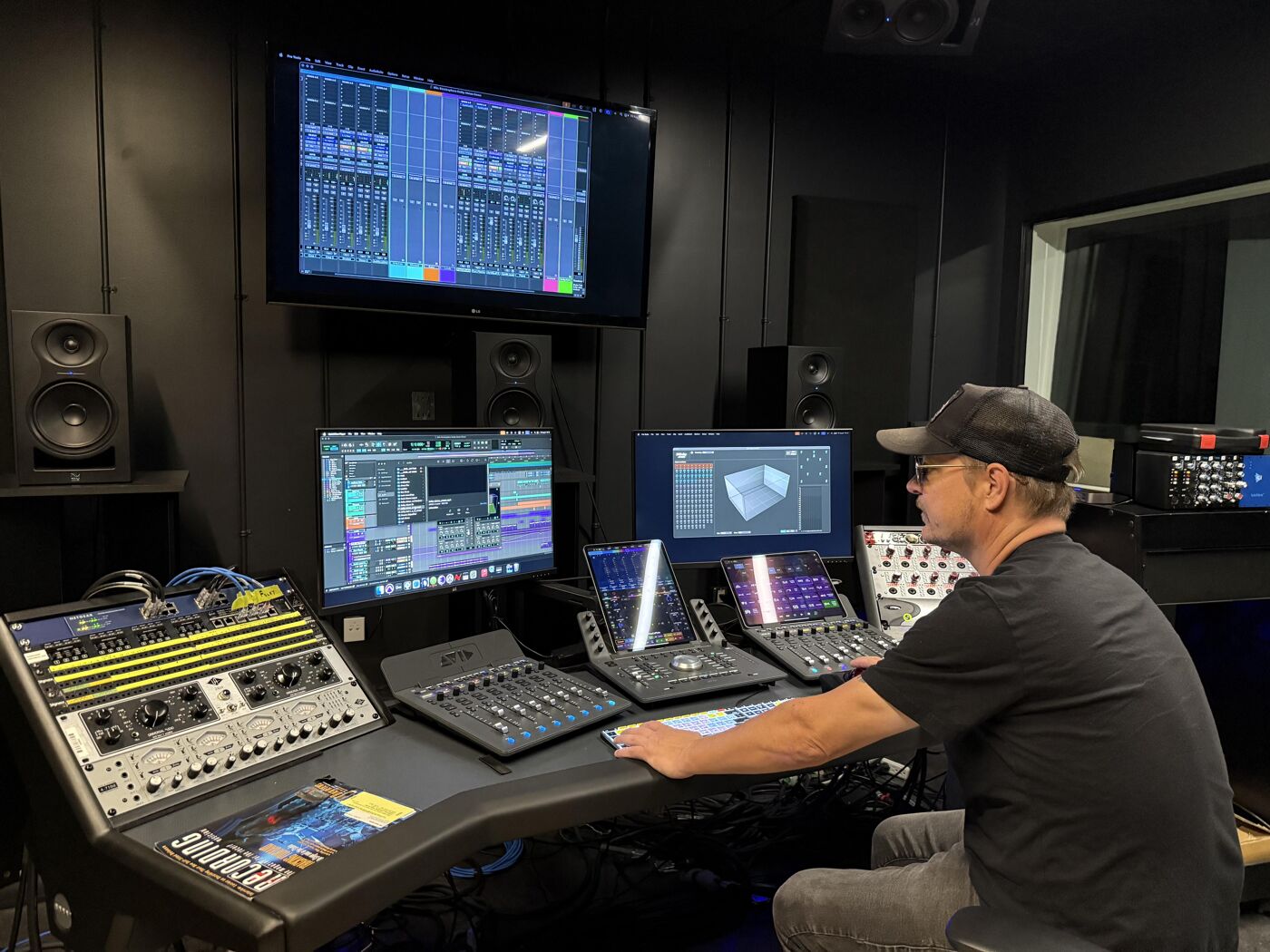 A man sits in a recording studio, surrounded by audio equipment including a mixing console, monitors displaying audio waveforms, and various control panels. He is focused on his work, likely mixing or mastering audio tracks.