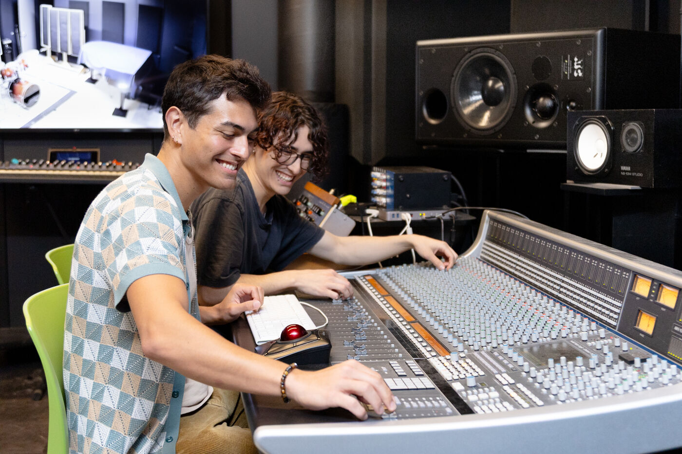 Two men smile as they work together at an audio mixing console in a recording studio, surrounded by professional equipment.