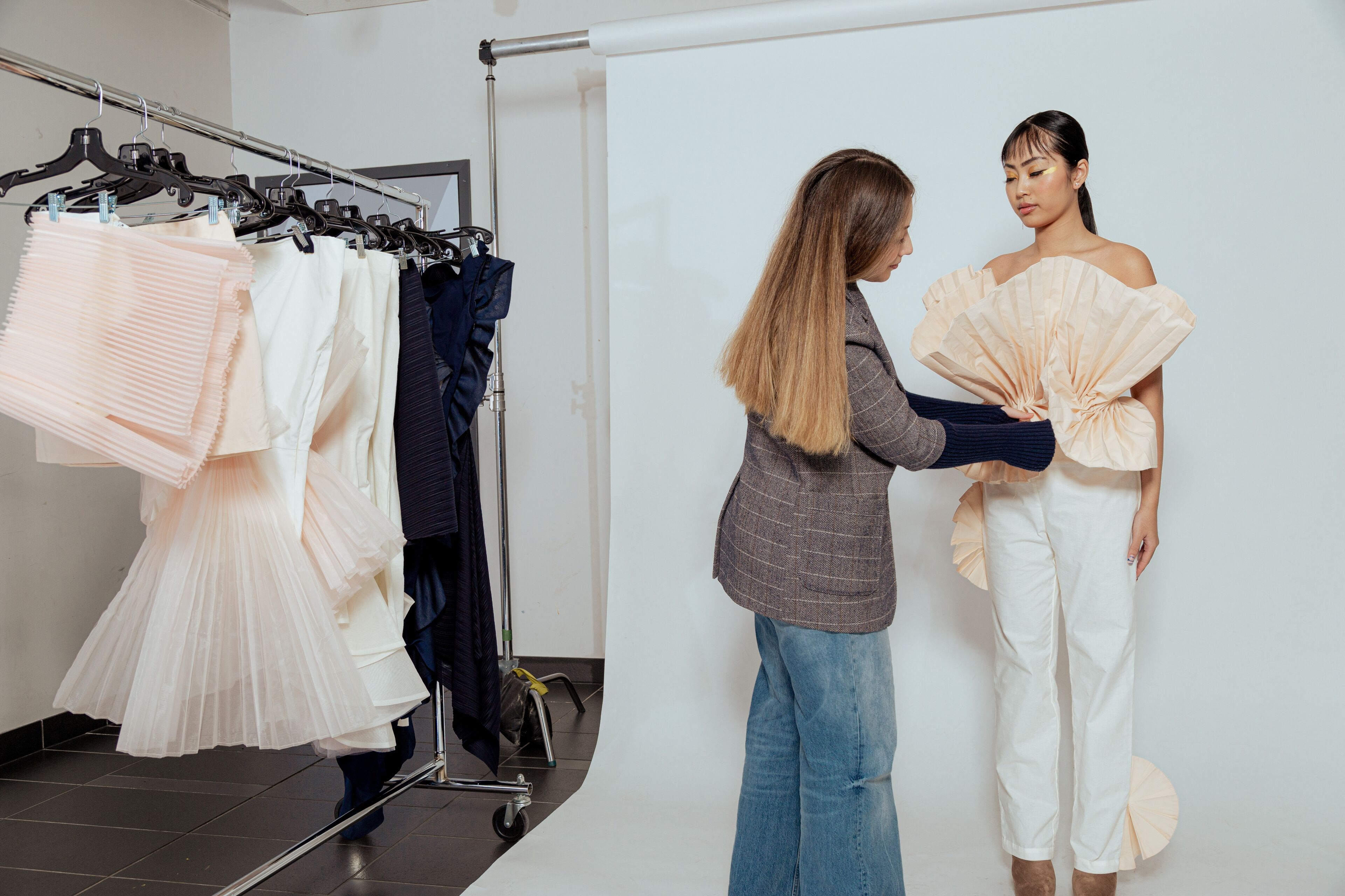 A designer adjusts a unique ruffled garment on a model in a fashion studio, with a rack of clothes in the background.