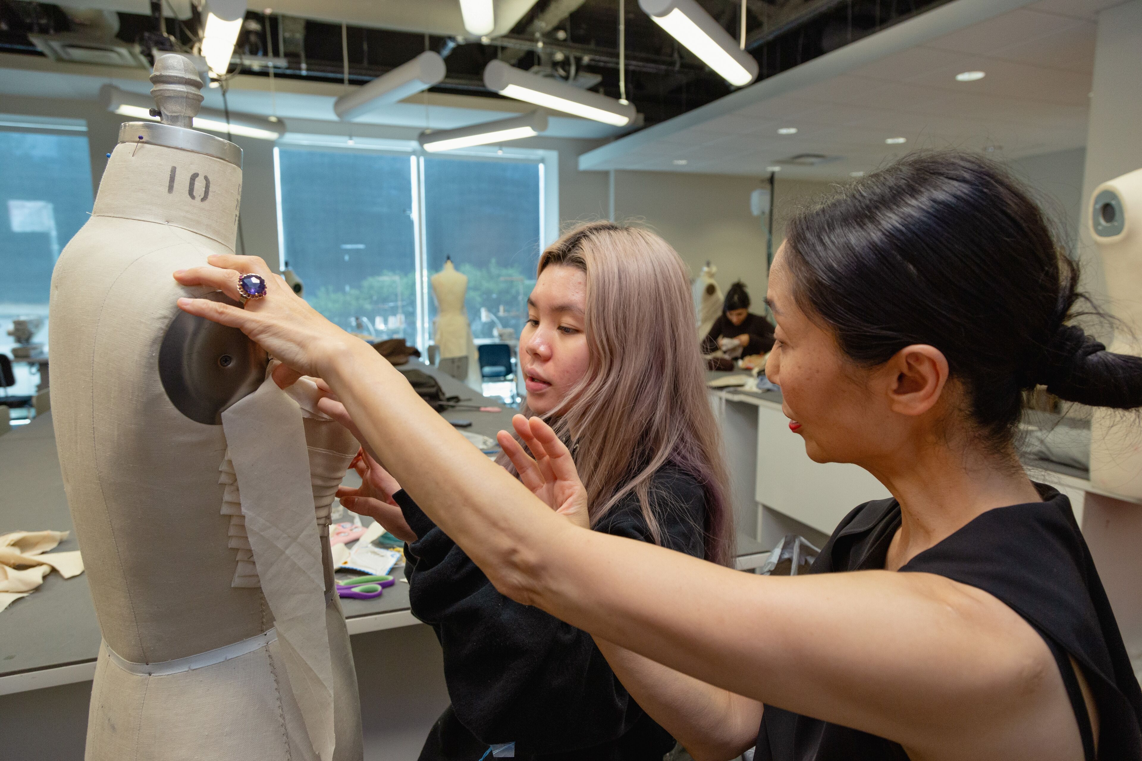 An experienced tailor instructs a young designer on dress fitting techniques on a mannequin.