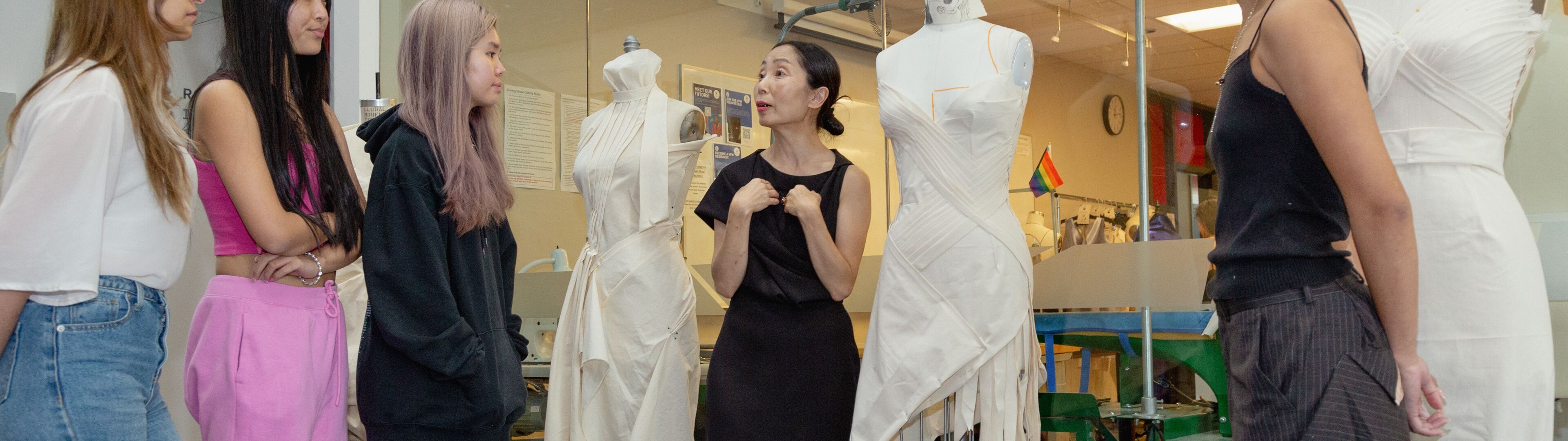 Students attentively listen as their instructor explains garment construction techniques beside mannequins.