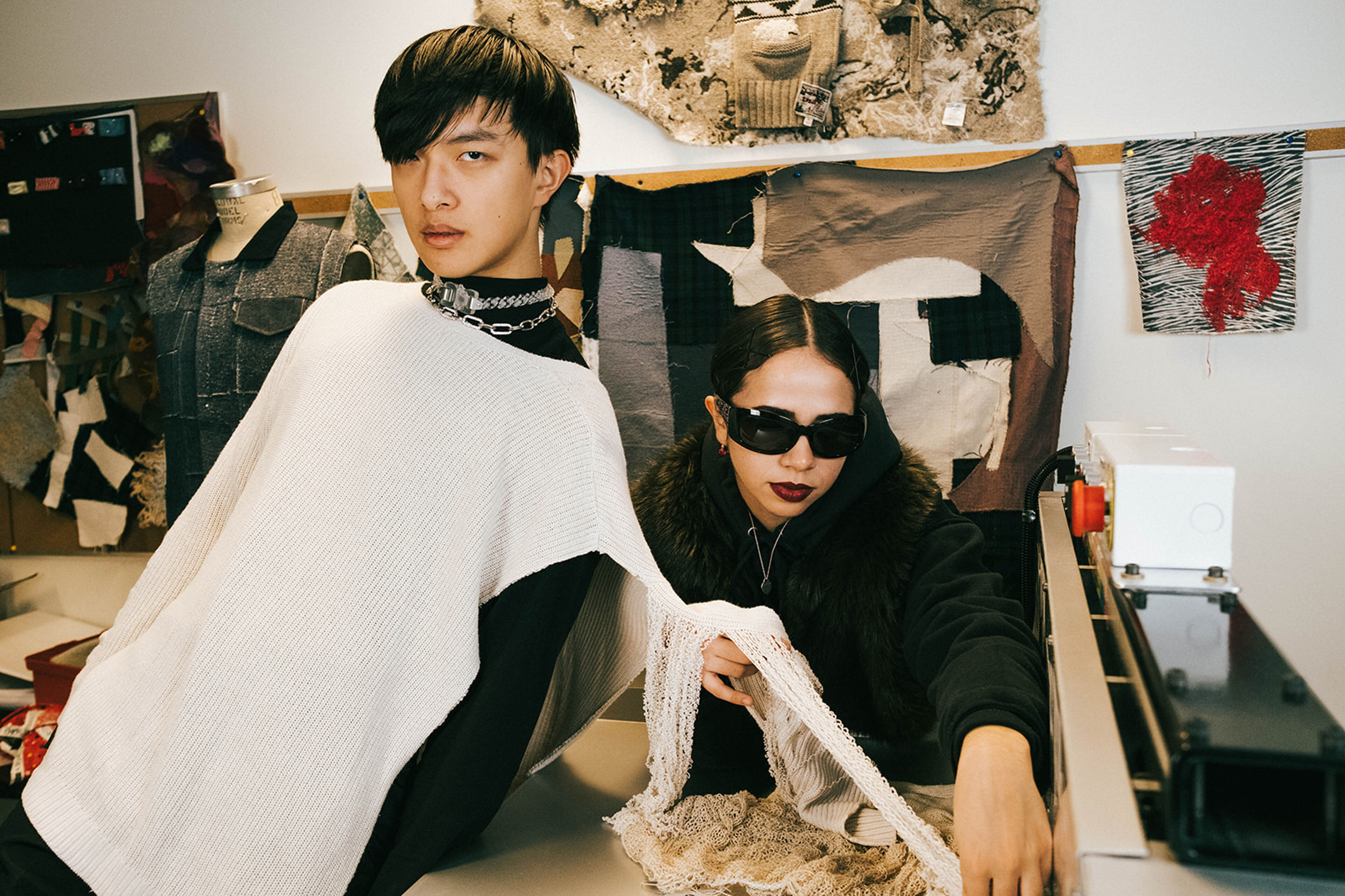 Two fashion designers in a creative studio, with textile samples and a sewing machine in the background.