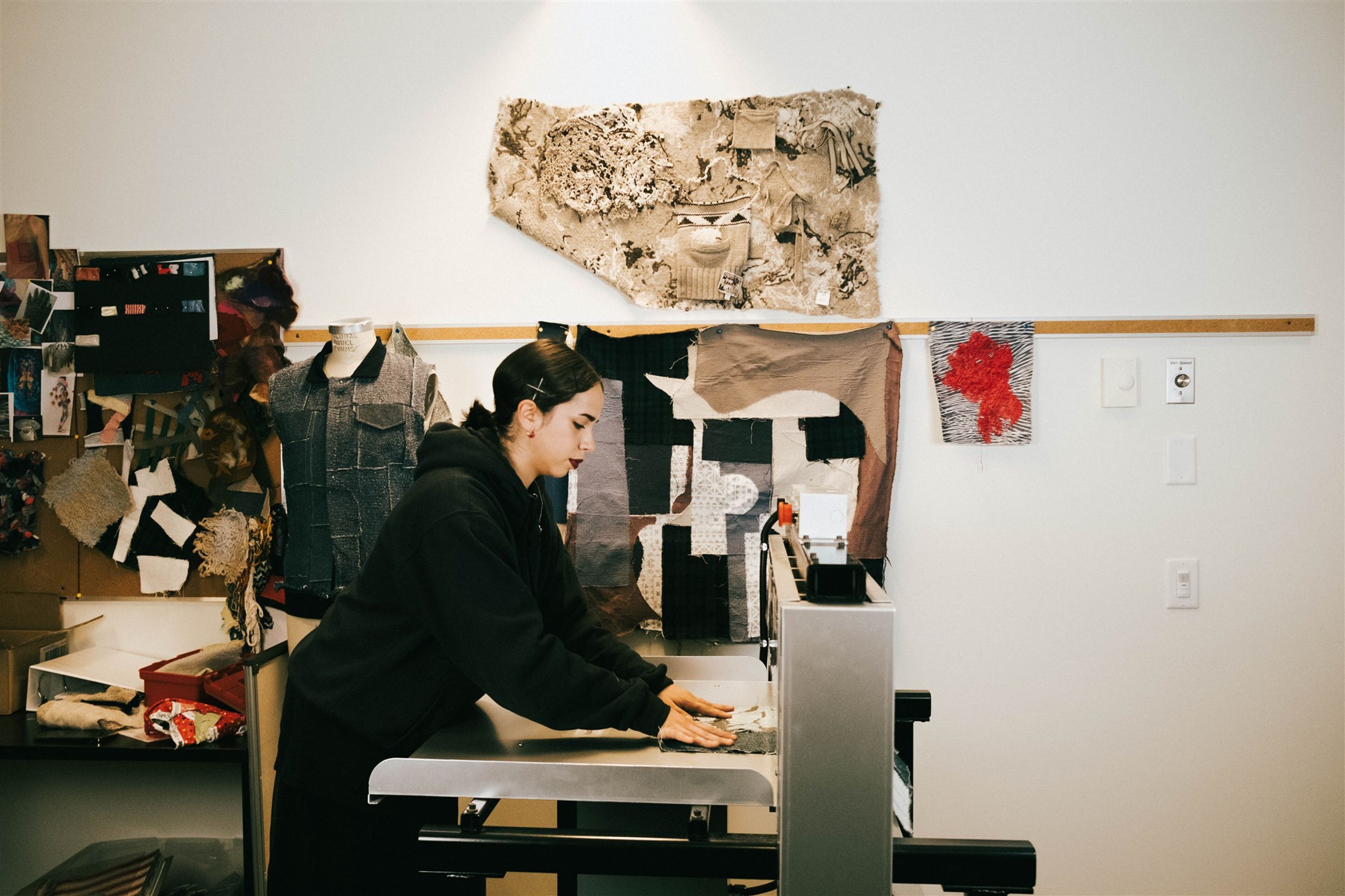 A focused fashion designer operates a sewing machine amidst a creative studio setting, surrounded by fabric samples and a mannequin adorned with a garment.
