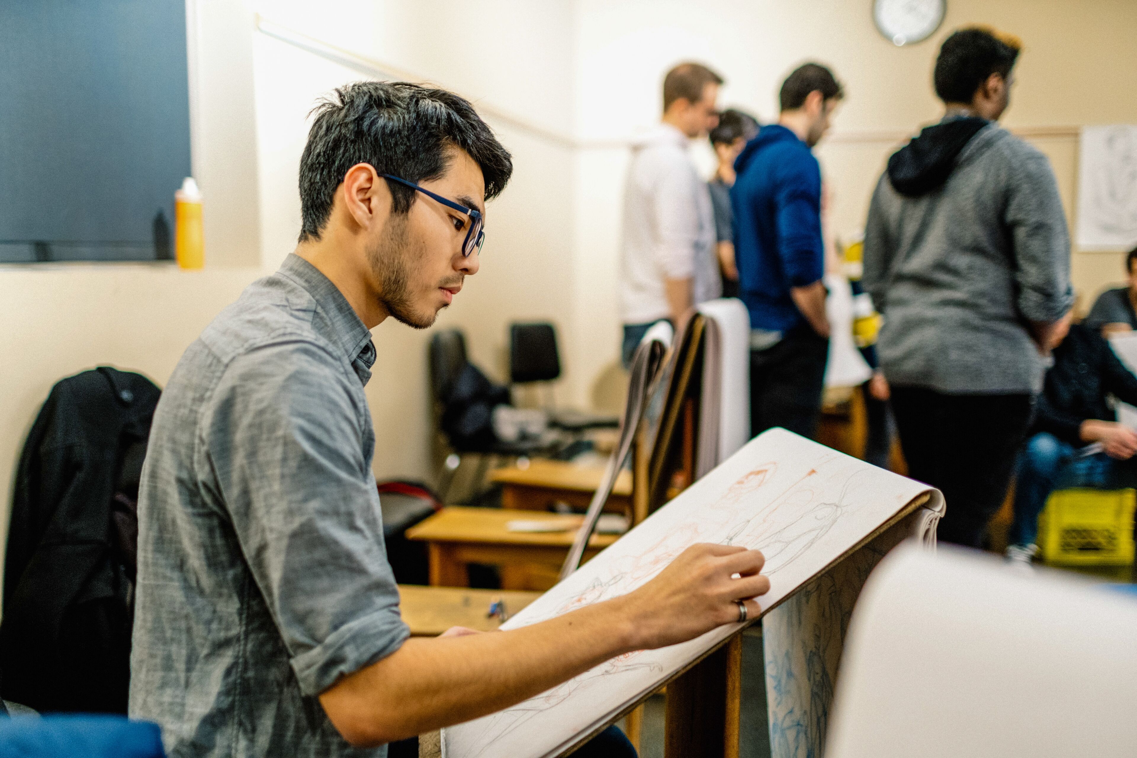 An artist intently sketches on a drawing board, immersed in a bustling classroom environment.