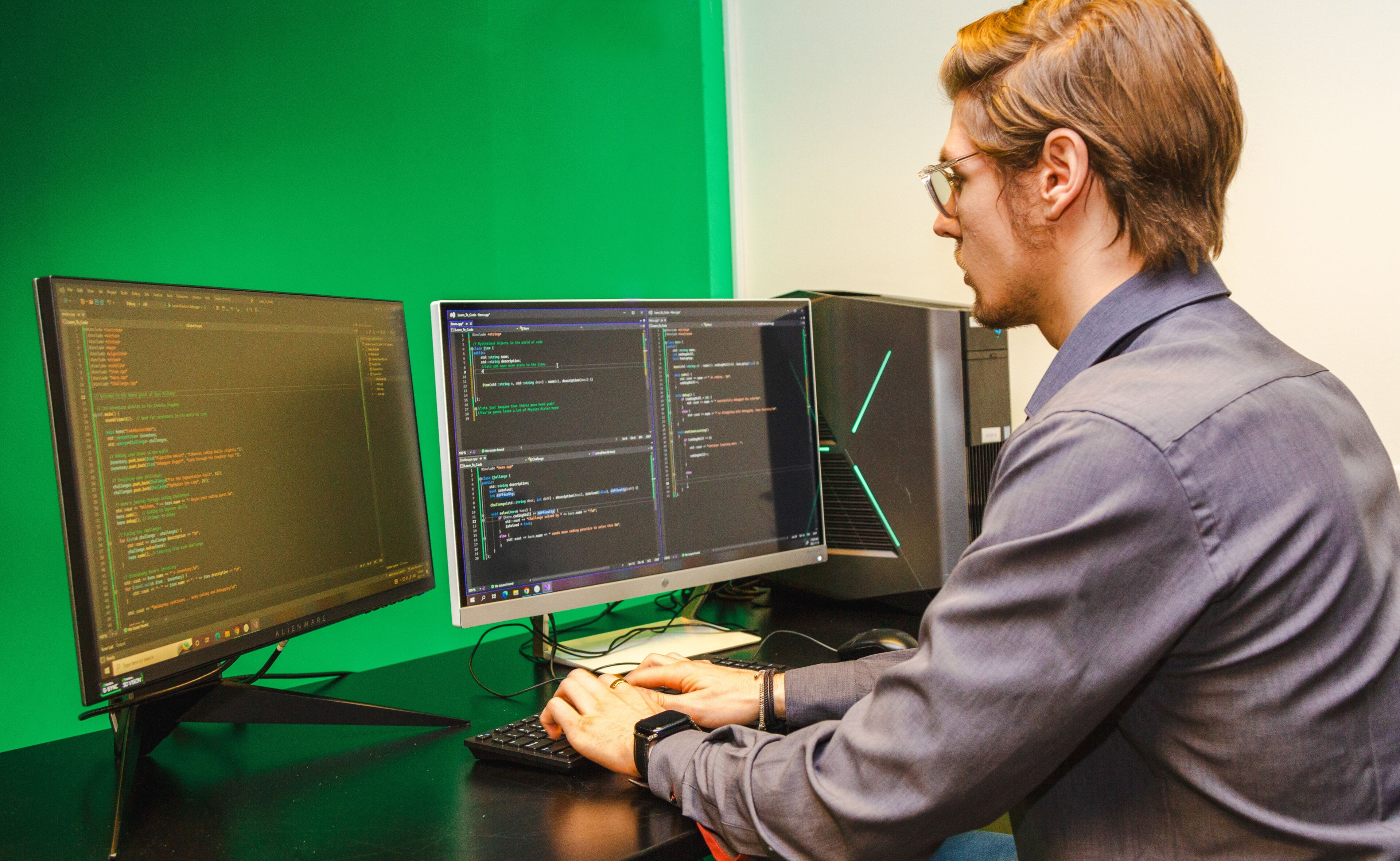 A focused male software developer works on code across dual monitors in an office with a green wall.