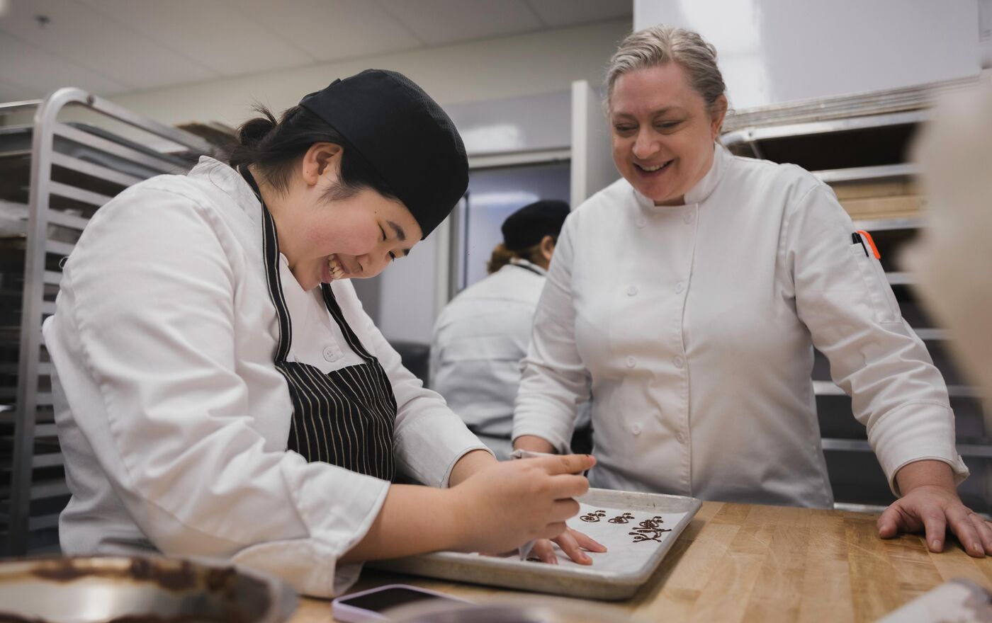 The image shows two chefs in a kitchen, one is working on a tray of food while the other is watching and smiling. They appear to be enjoying their work.