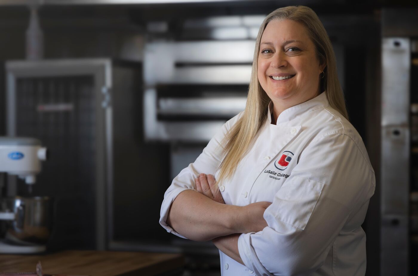 The image shows a smiling woman in a chef's coat with her arms crossed, standing in a kitchen. There is a mixer and oven in the background.