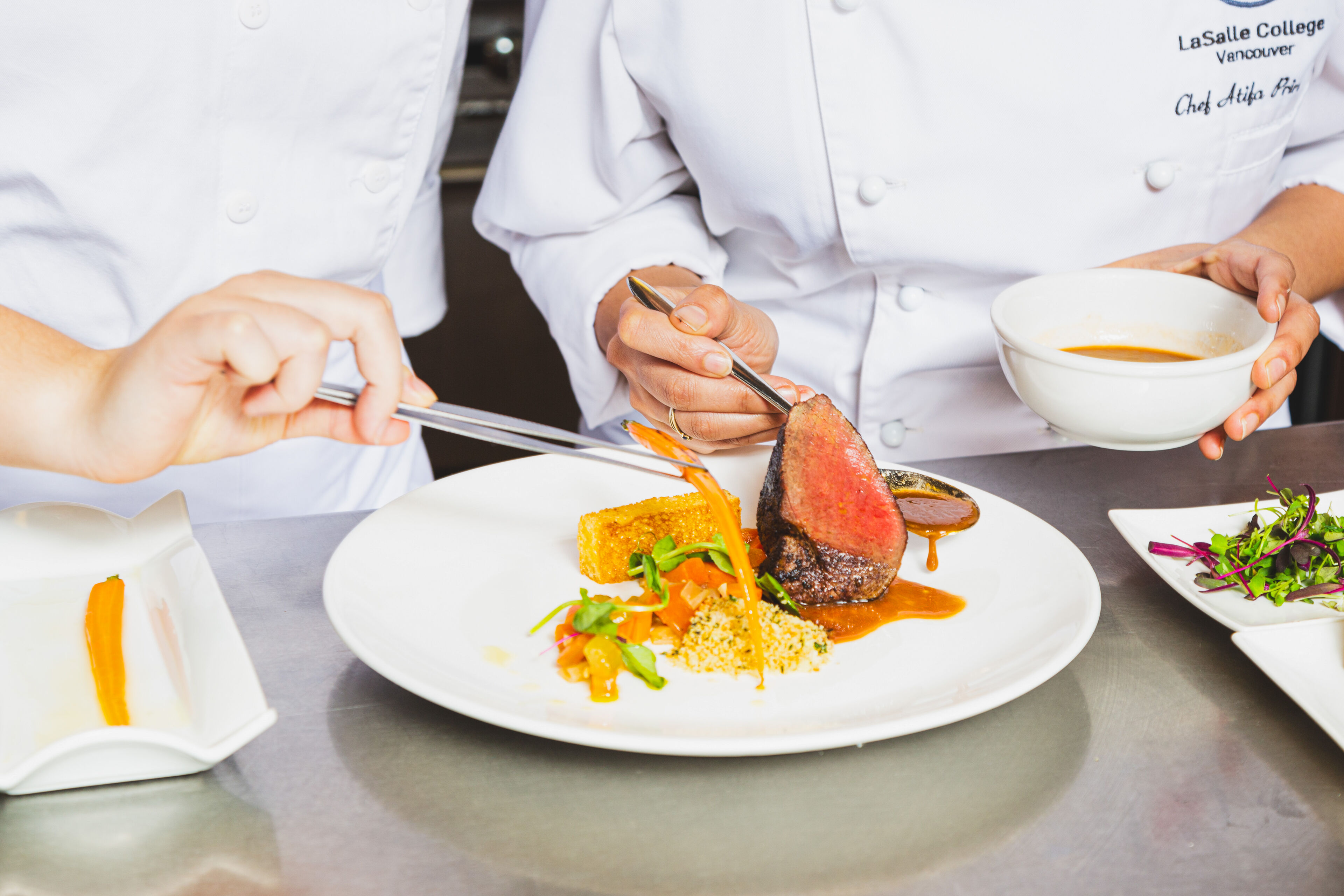 Chef garnishing a medium-rare beef steak with sauce, served with grains and vegetables, in a professional kitchen.