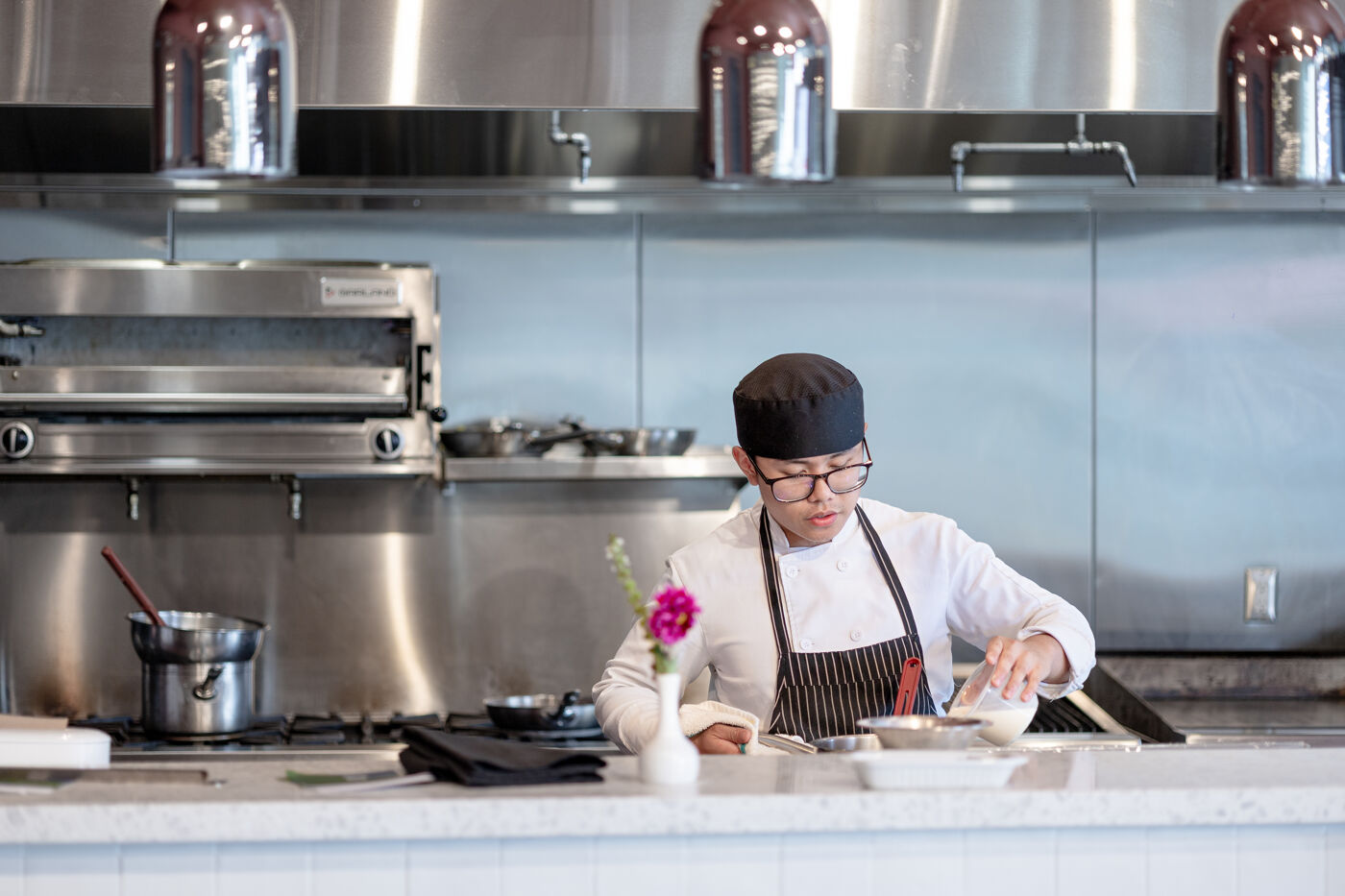 A chef wearing glasses and a black cap is cooking in a professional kitchen, focused on their task with ingredients laid out in front of them.