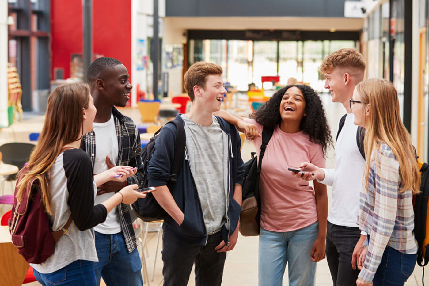 The image shows a group of young people, including both men and women, standing together in a hallway or a room. They are engaged in conversation and smiling, which suggests that they are enjoying each other's company. Some of them are holding cell phones, which might be used for communication, sharing photos, or browsing the internet. The group appears to be friends or acquaintances, and they seem to be having a good time together.