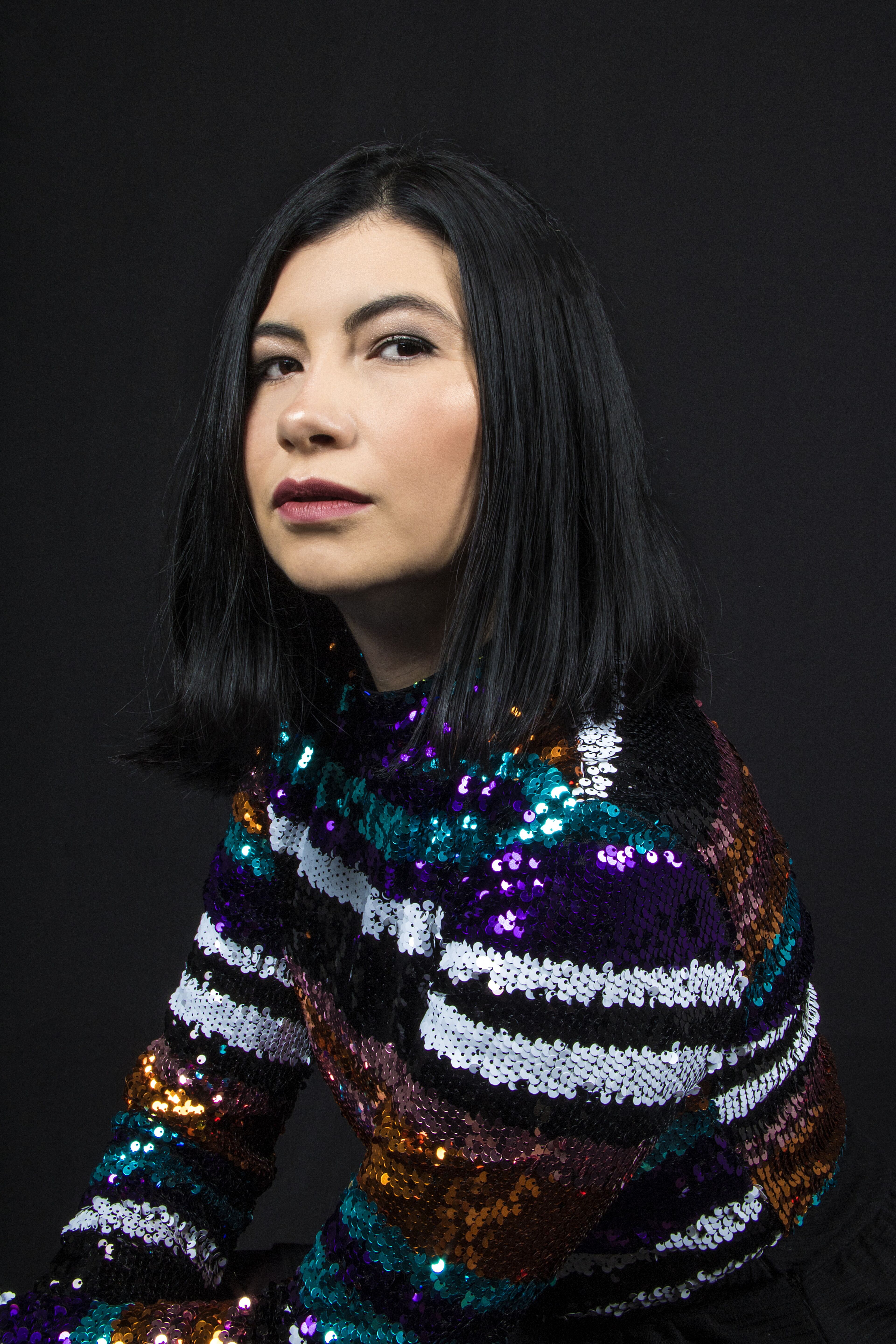 A poised woman with black hair against a black background, wearing a multicolored sequined top.