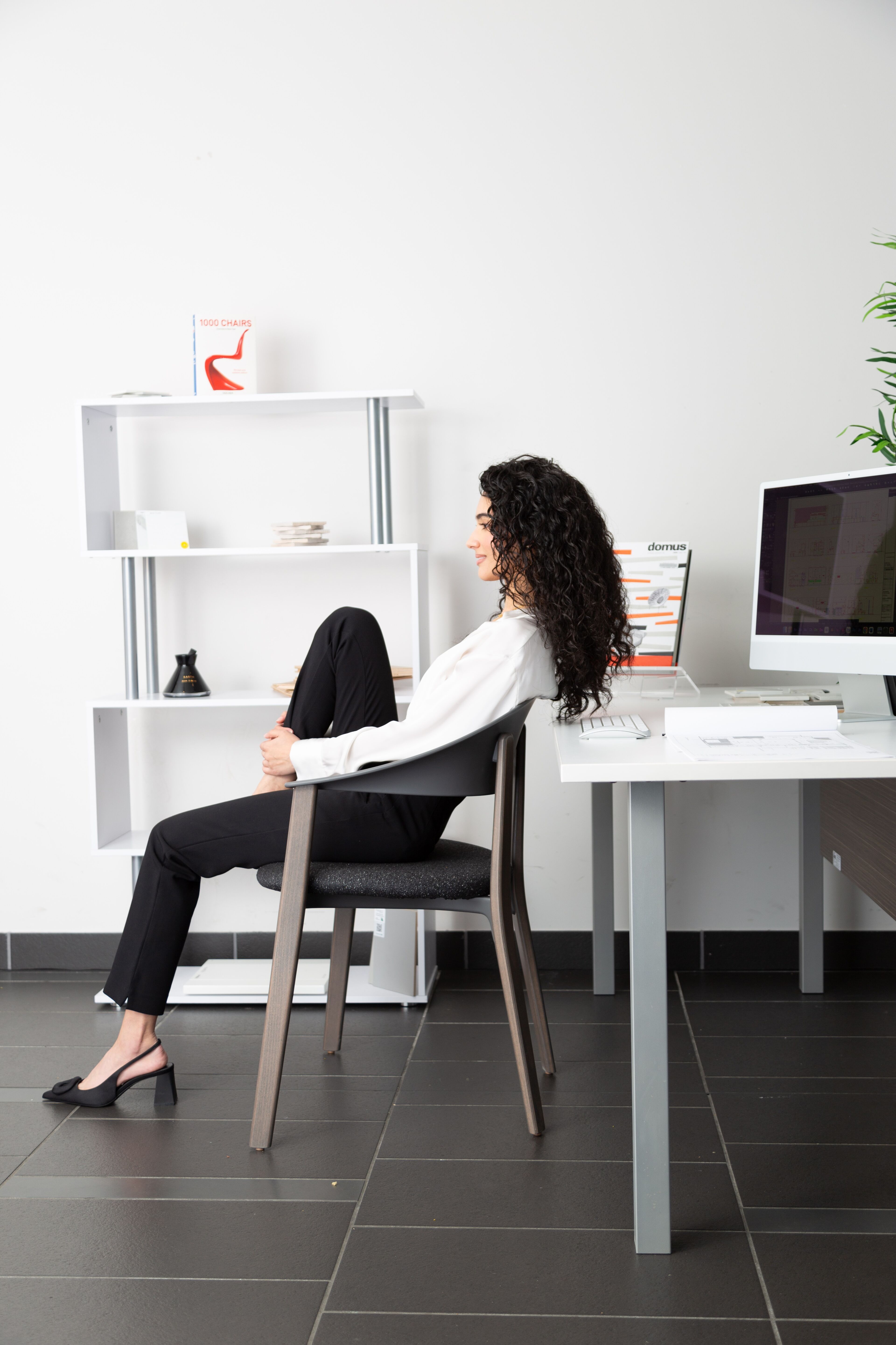 A professional woman sits sideways on a chair at a minimalist office, facing a white shelf and looking away from a computer screen.