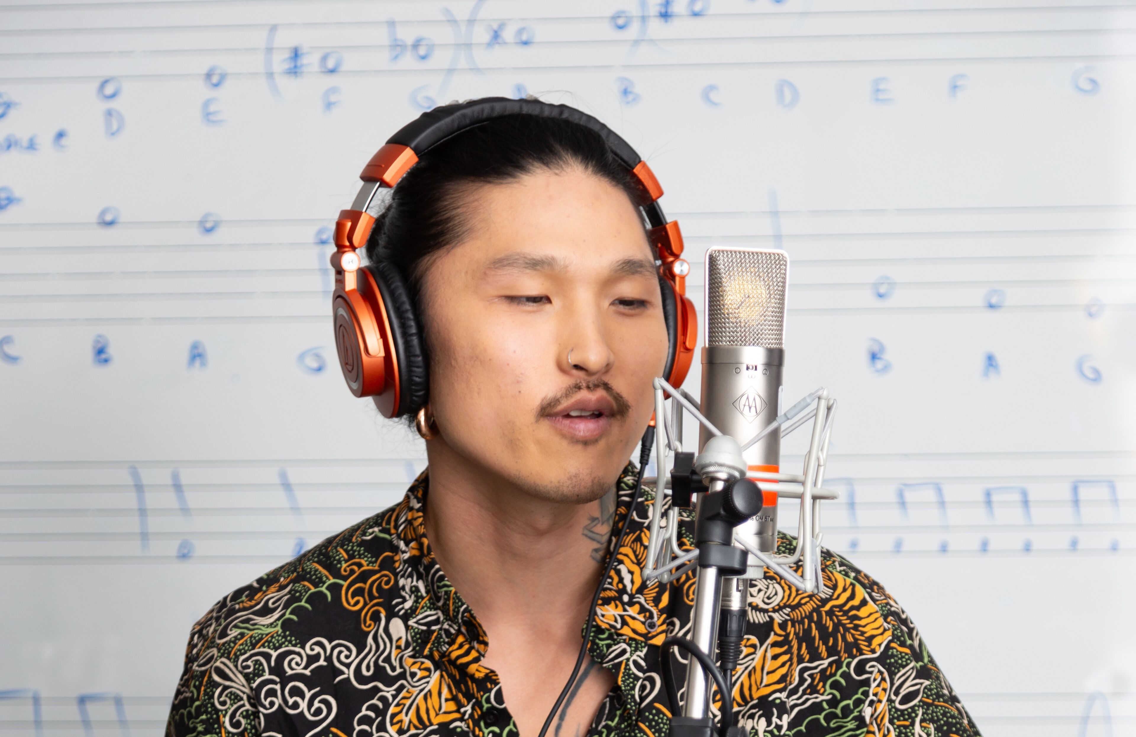 A tattooed man in an ornate shirt singing into a studio microphone with headphones.