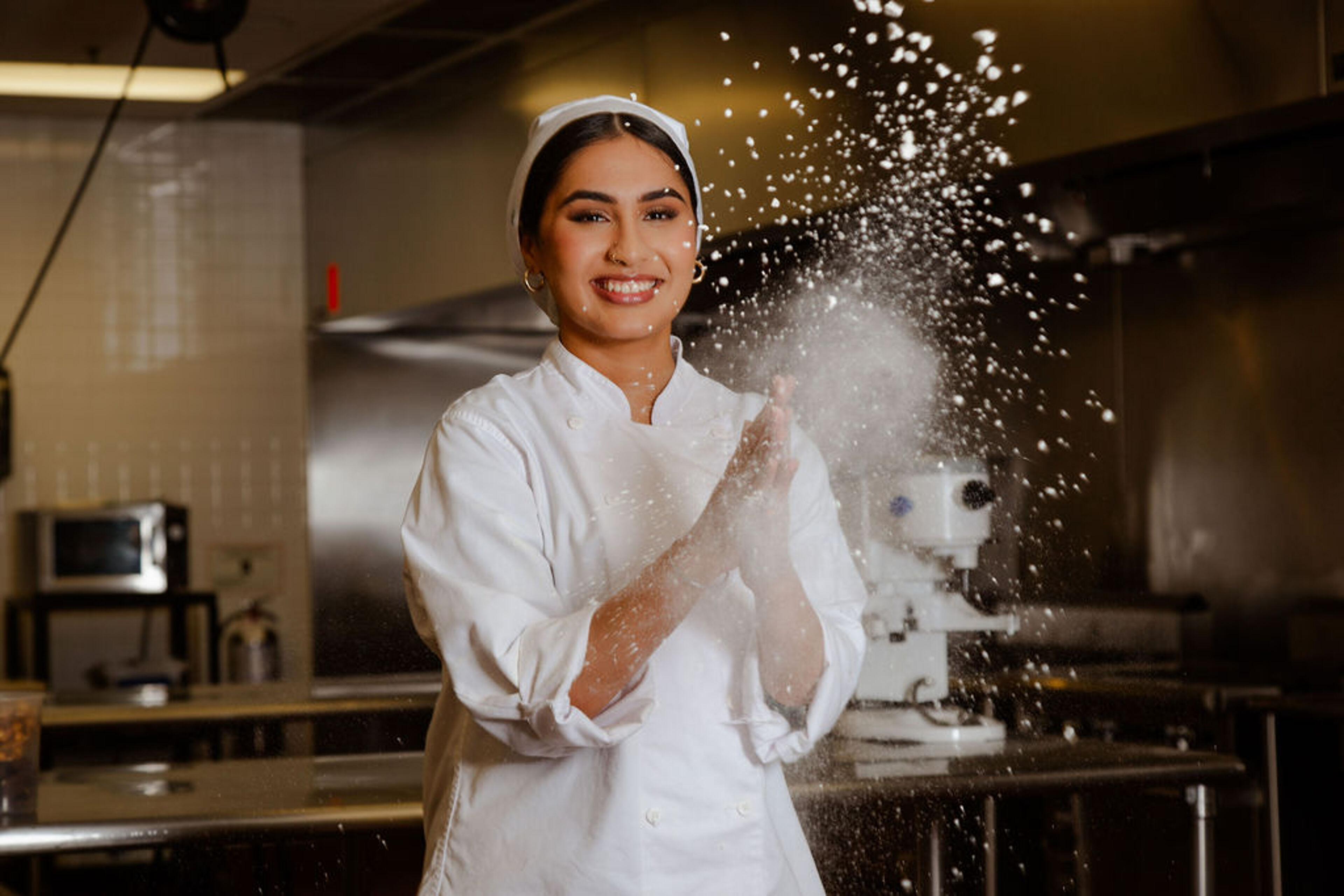 A smiling female chef clapping her hands with flour in a professional kitchen, creating a dynamic cloud of flour around her.