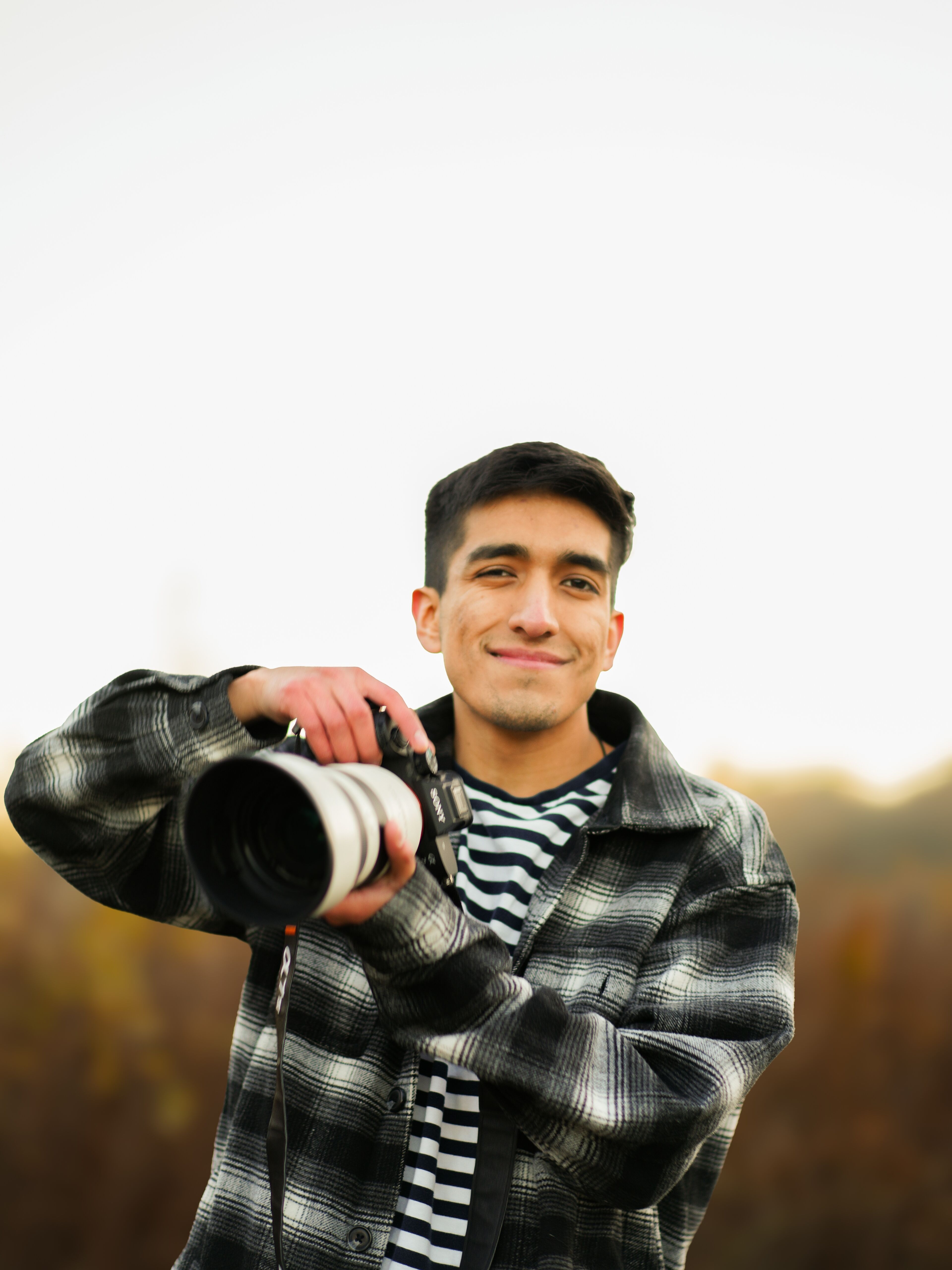 A cheerful young man holding a professional camera in an autumnal outdoor setting.
