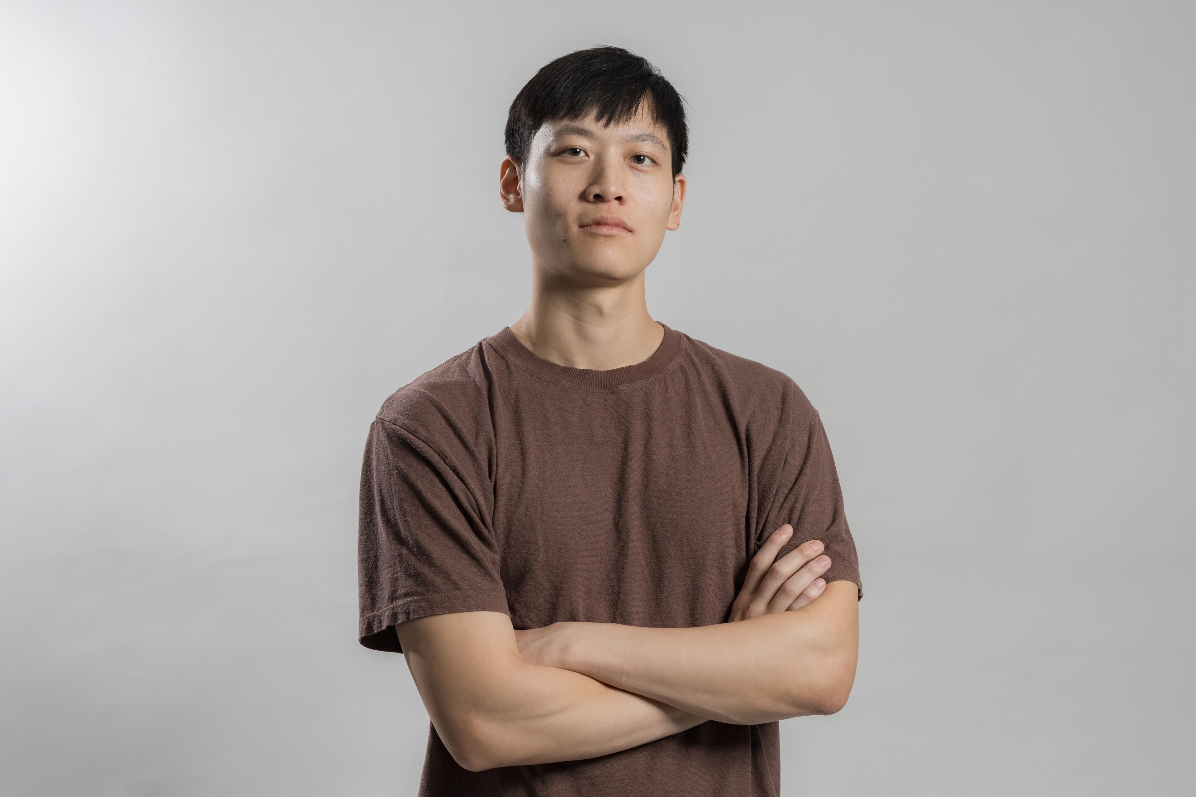 A young adult male stands with arms crossed, exuding confidence, against a neutral grey background.