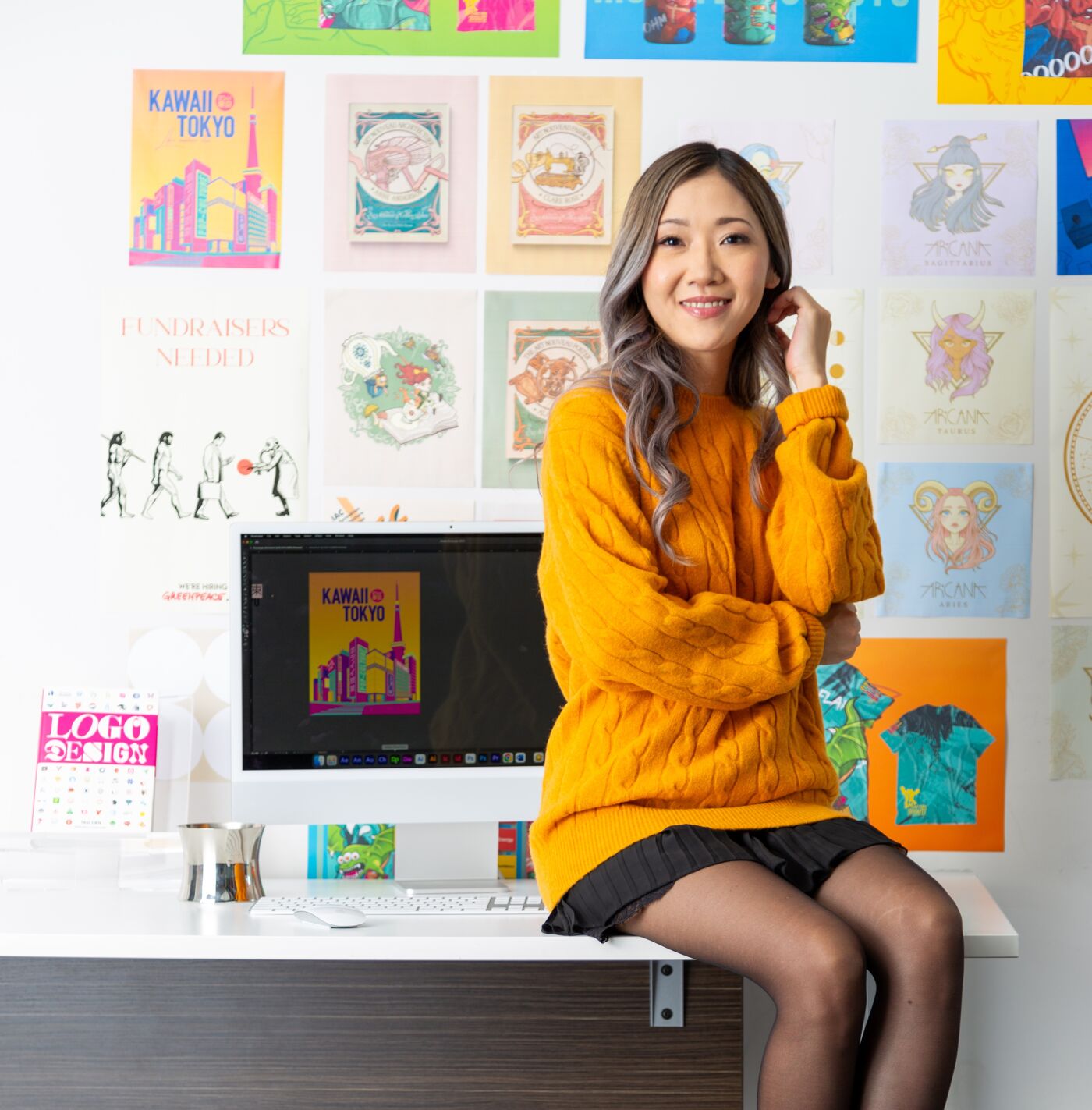 A smiling woman in a mustard sweater and skirt sits at a desk adorned with colorful art and design paraphernalia.