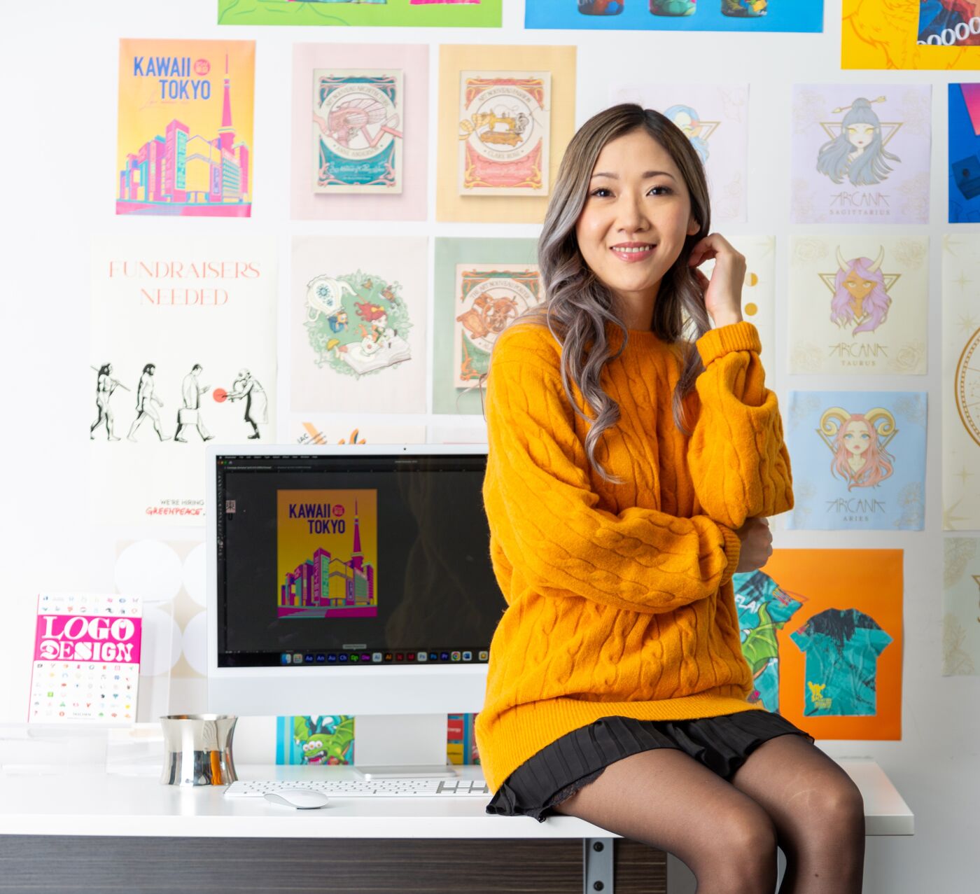 A smiling woman in a mustard sweater and skirt sits at a desk adorned with colorful art and design paraphernalia.