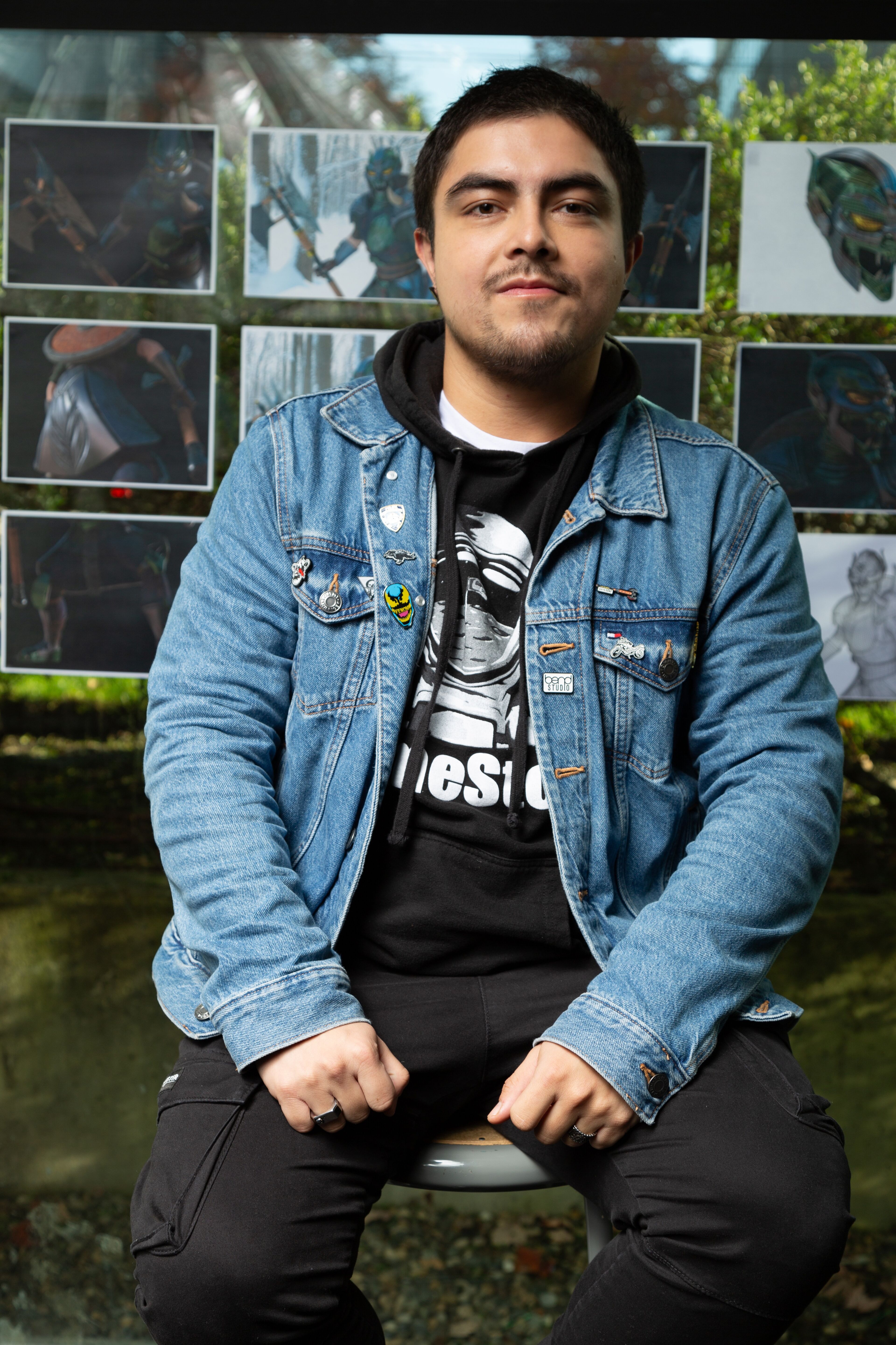 A confident male artist seated before his character sketches, sporting a denim jacket adorned with pins.