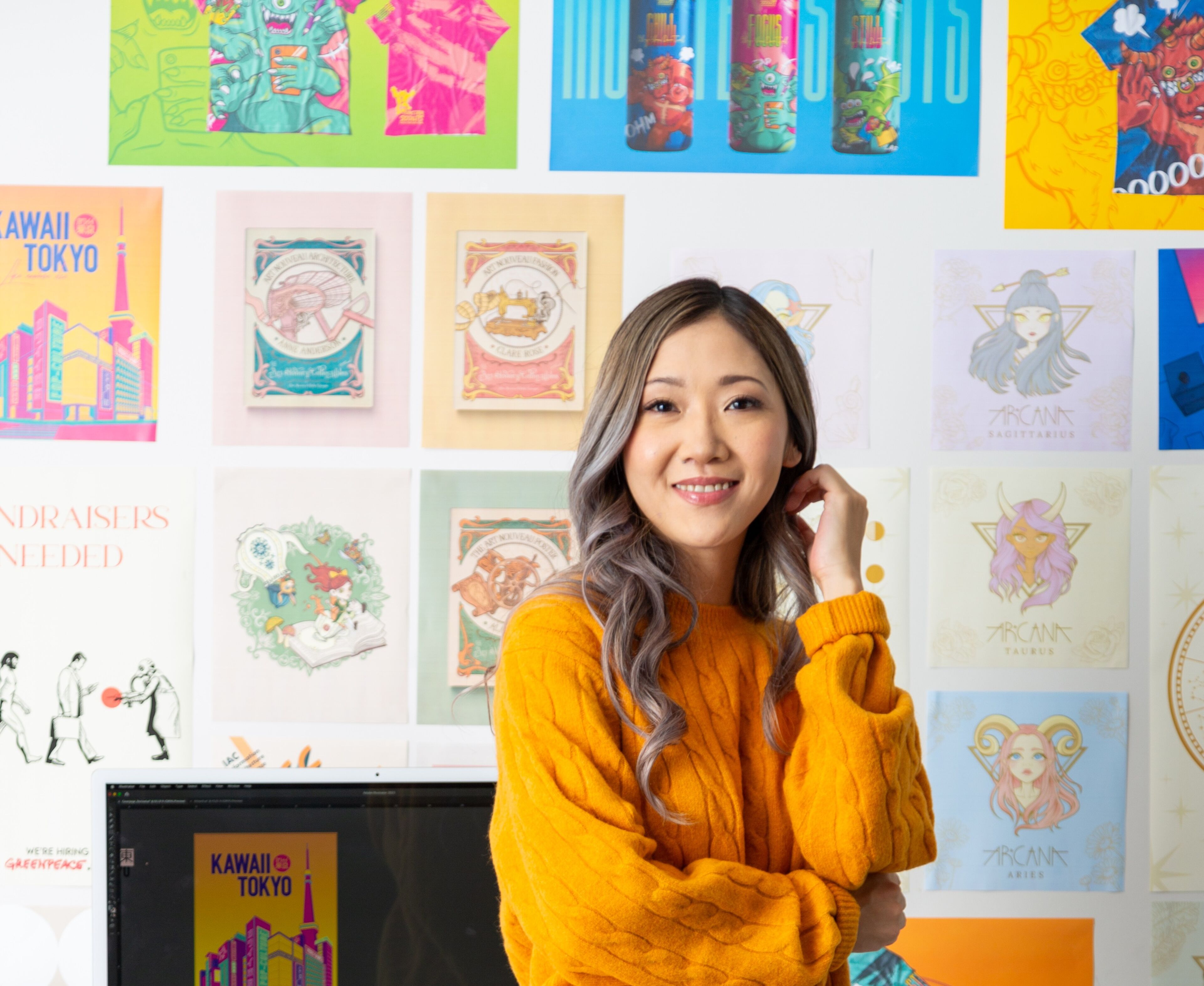 A smiling woman in a mustard sweater and skirt sits at a desk adorned with colorful art and design paraphernalia.