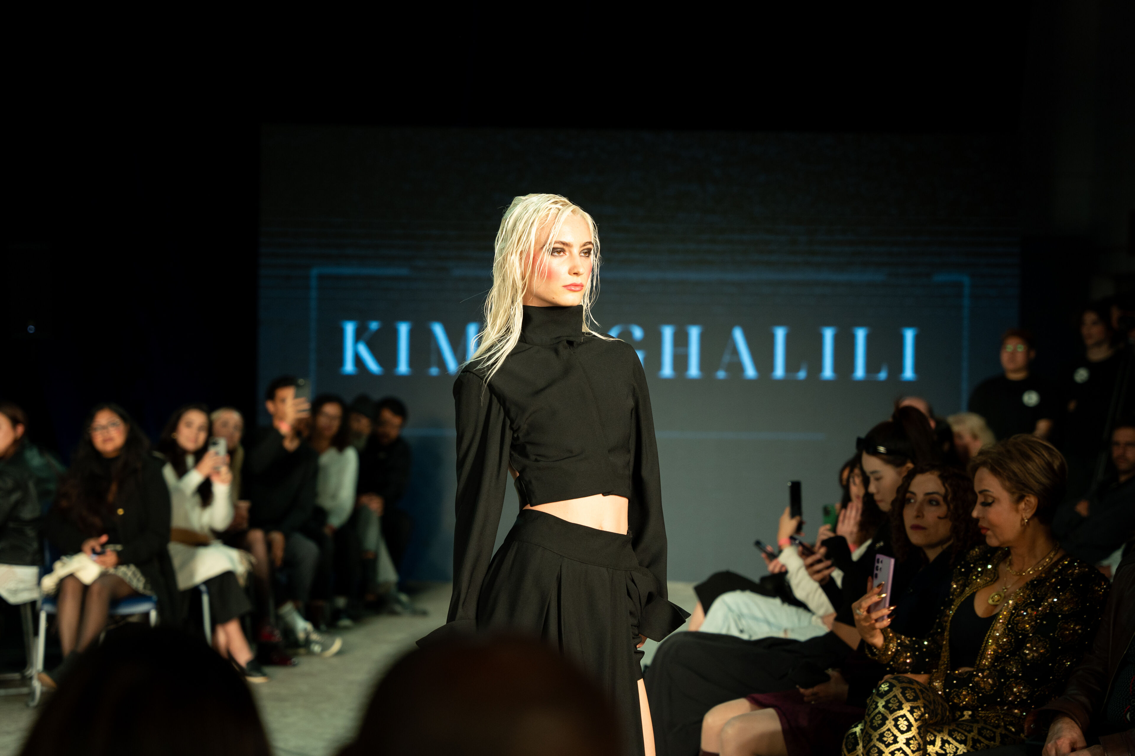 Model showcasing a black crop top ensemble on the runway during a fashion show, with attentive audience in the background.