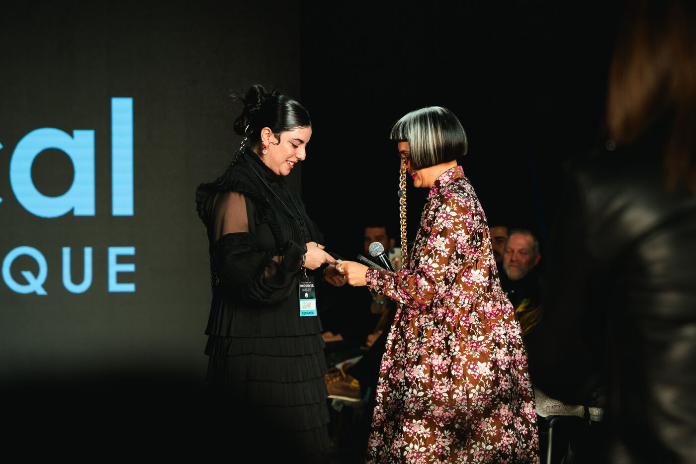Two women interacting at a fashion event, one hands a microphone to the other against a backdrop with the words "Digital Vogue".