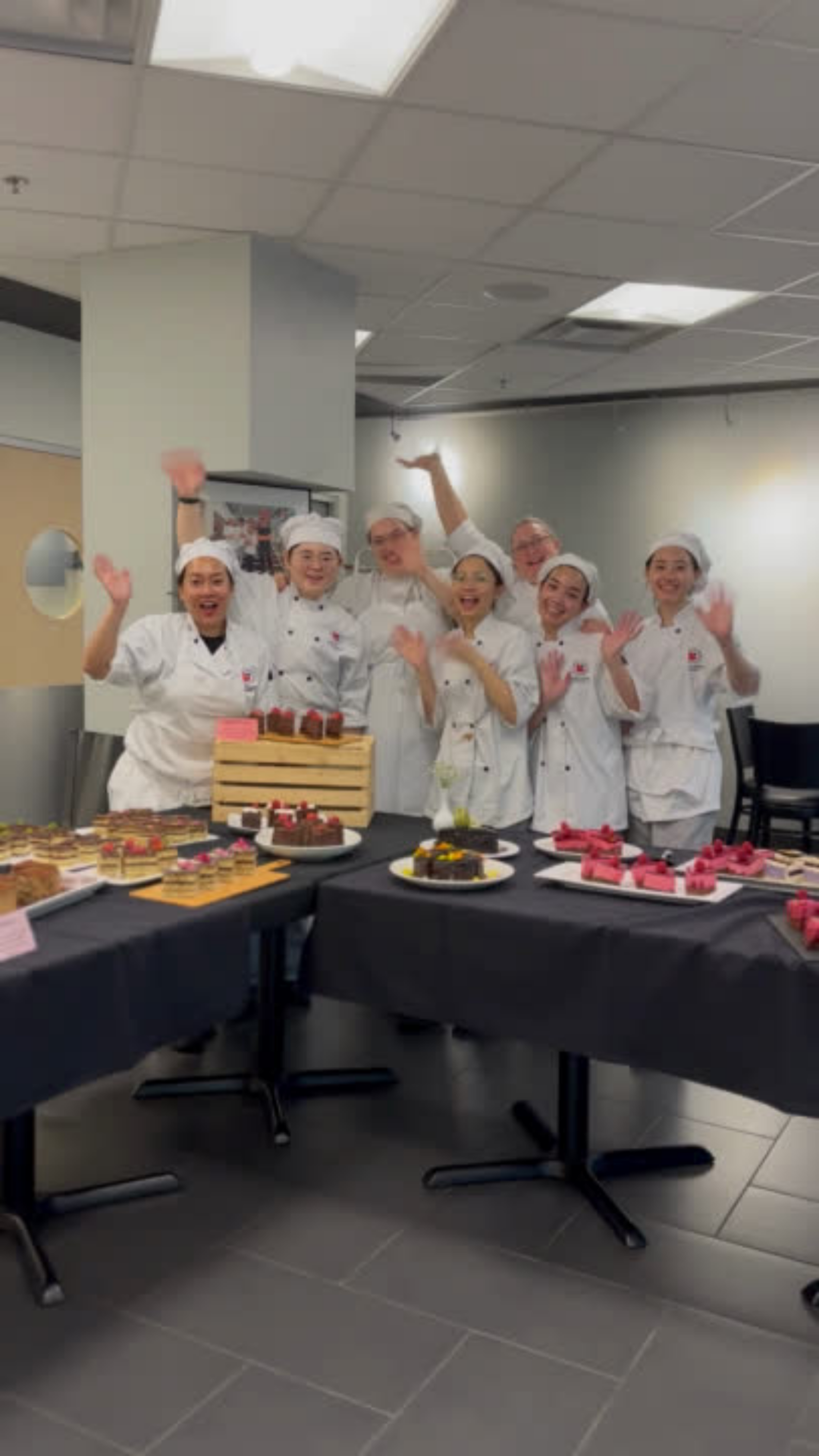 A team of chefs in white uniforms joyfully raise their hands, surrounded by a spread of elegant desserts.
