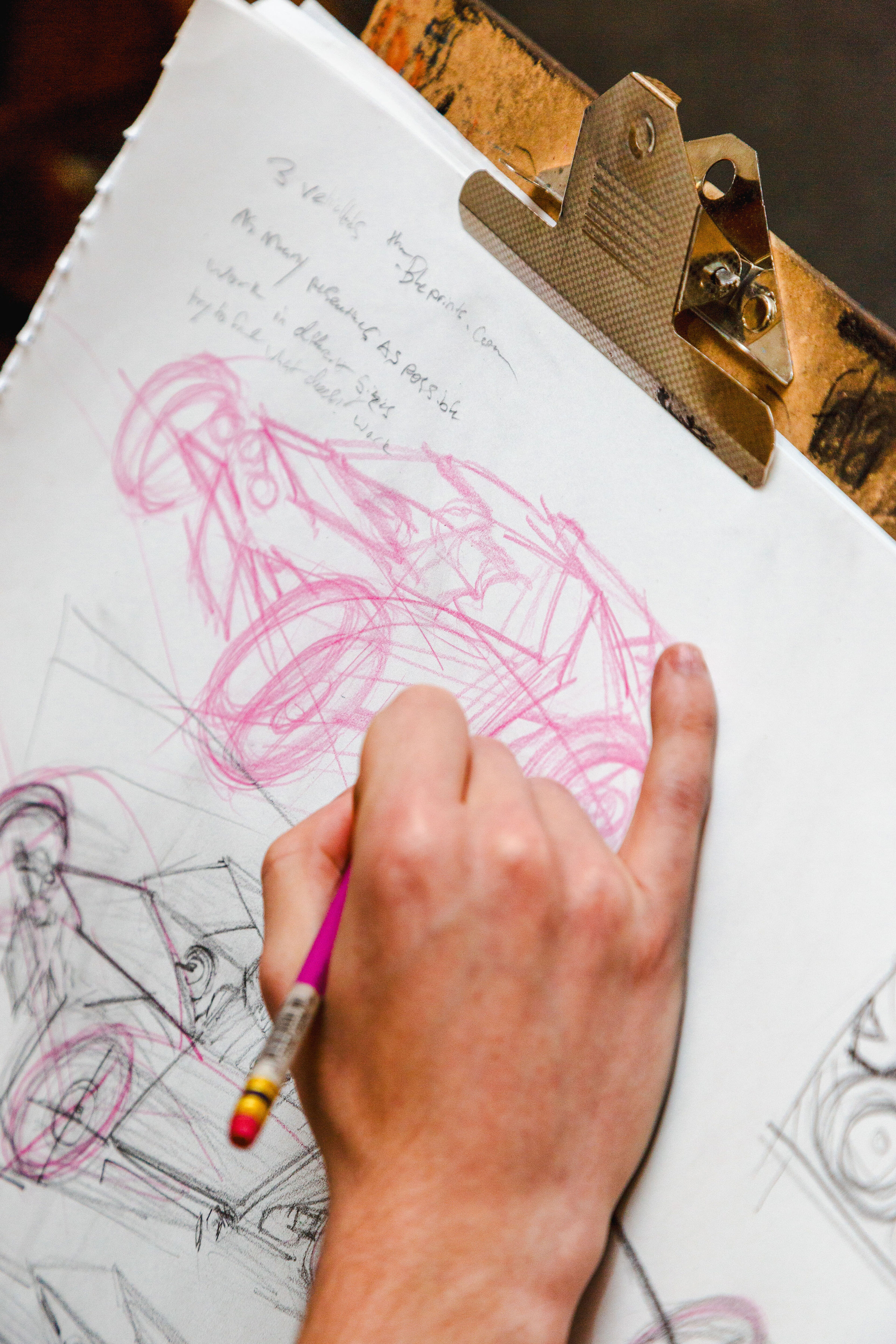 A close-up view of an artist's hand as they draw preliminary sketches with a red pencil on paper, clipped to a wooden clipboard.