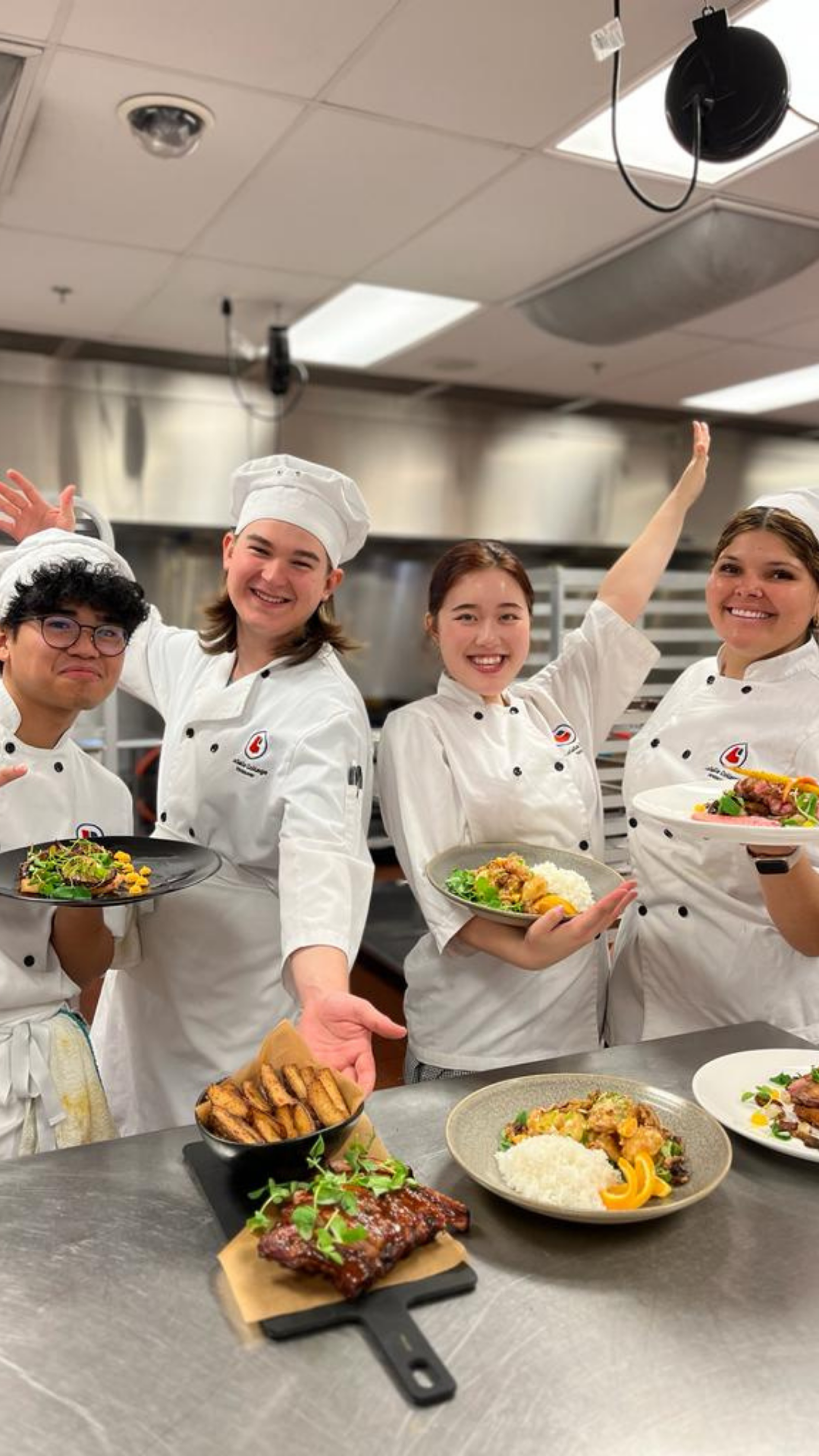 Four smiling culinary students in white chef uniforms proudly display an assortment of cooked dishes in a professional kitchen.