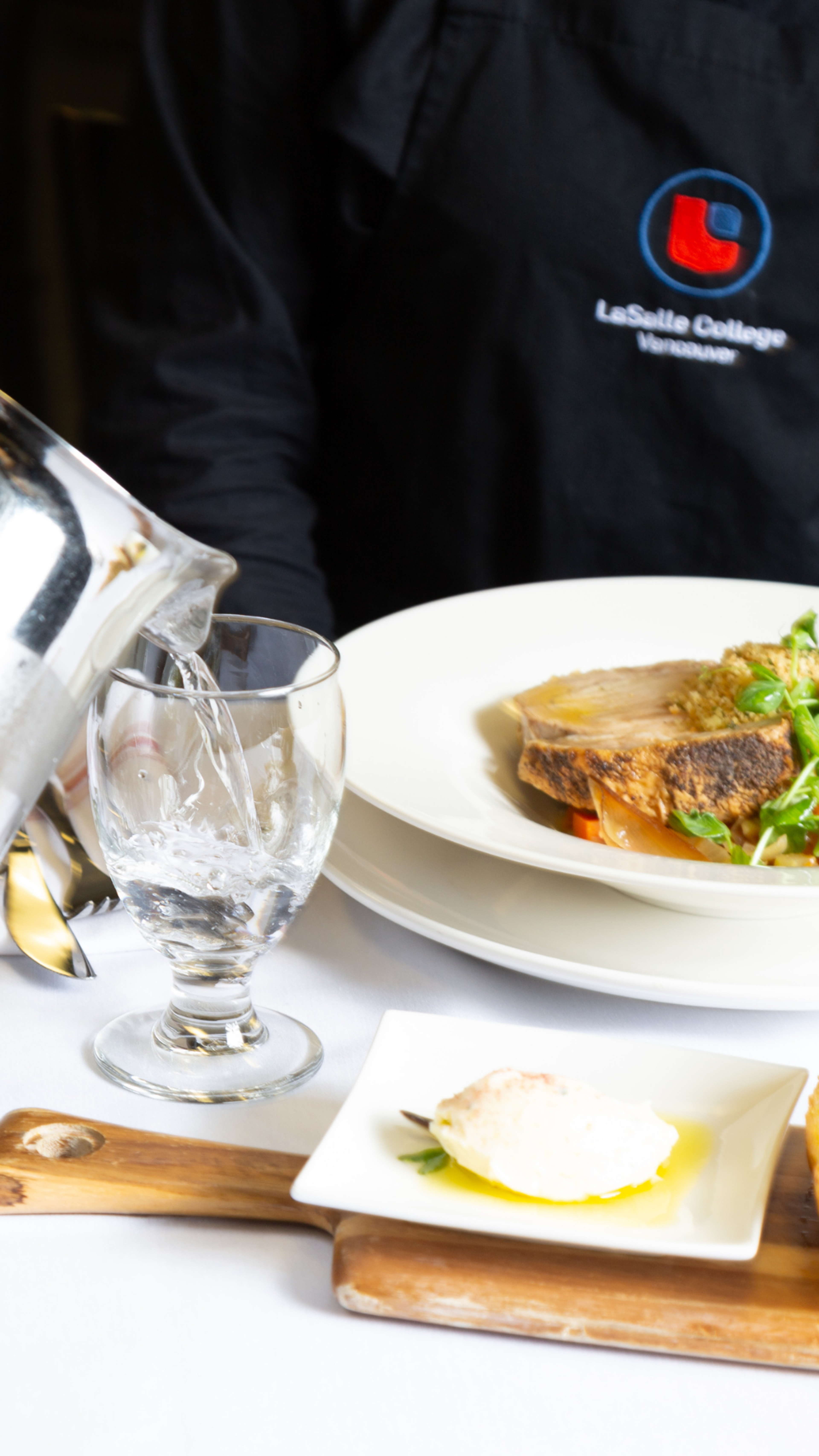 A server in a black apron, emblazoned with "LaSalle College Vancouver," pours water into a glass at a fine dining table featuring a dish of pan-seared fish and a side of creamy spread.
