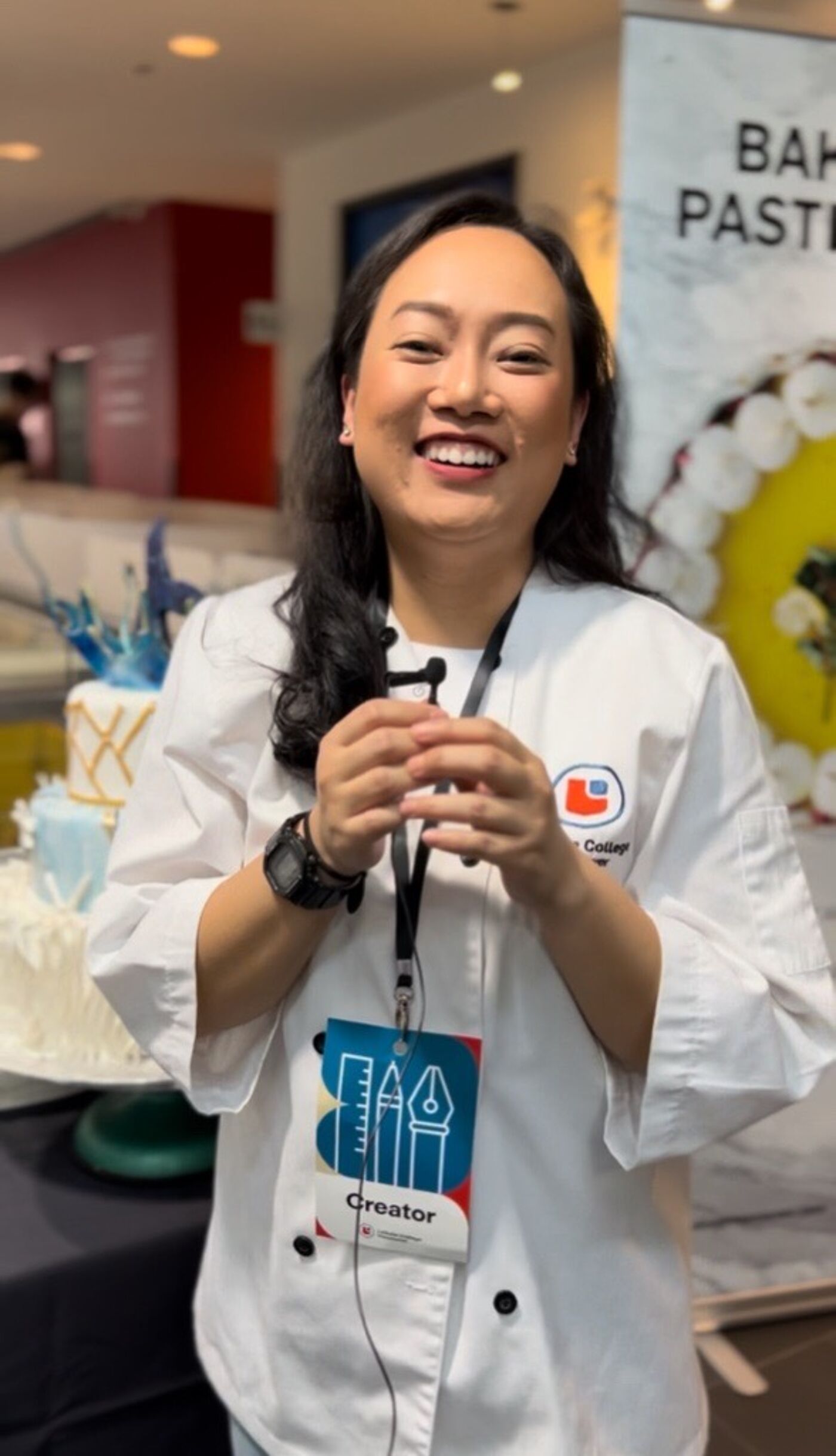 A smiling woman in a white chef's coat wears an ID badge that reads "Creator." She is surrounded by cakes and appears to be at a culinary event.