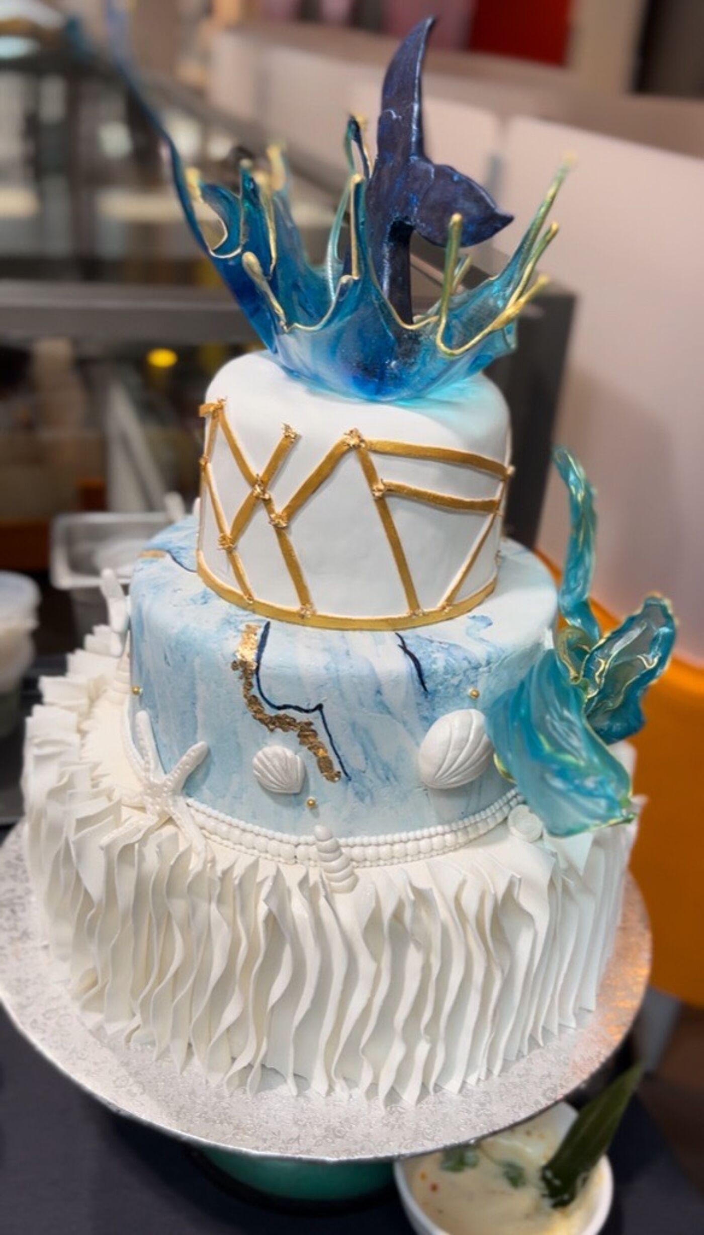 This three-tiered cake features an aquatic theme with whale tail and wave-like decorations made of blue candy. The bottom tier is adorned with white ruffles resembling sea foam.