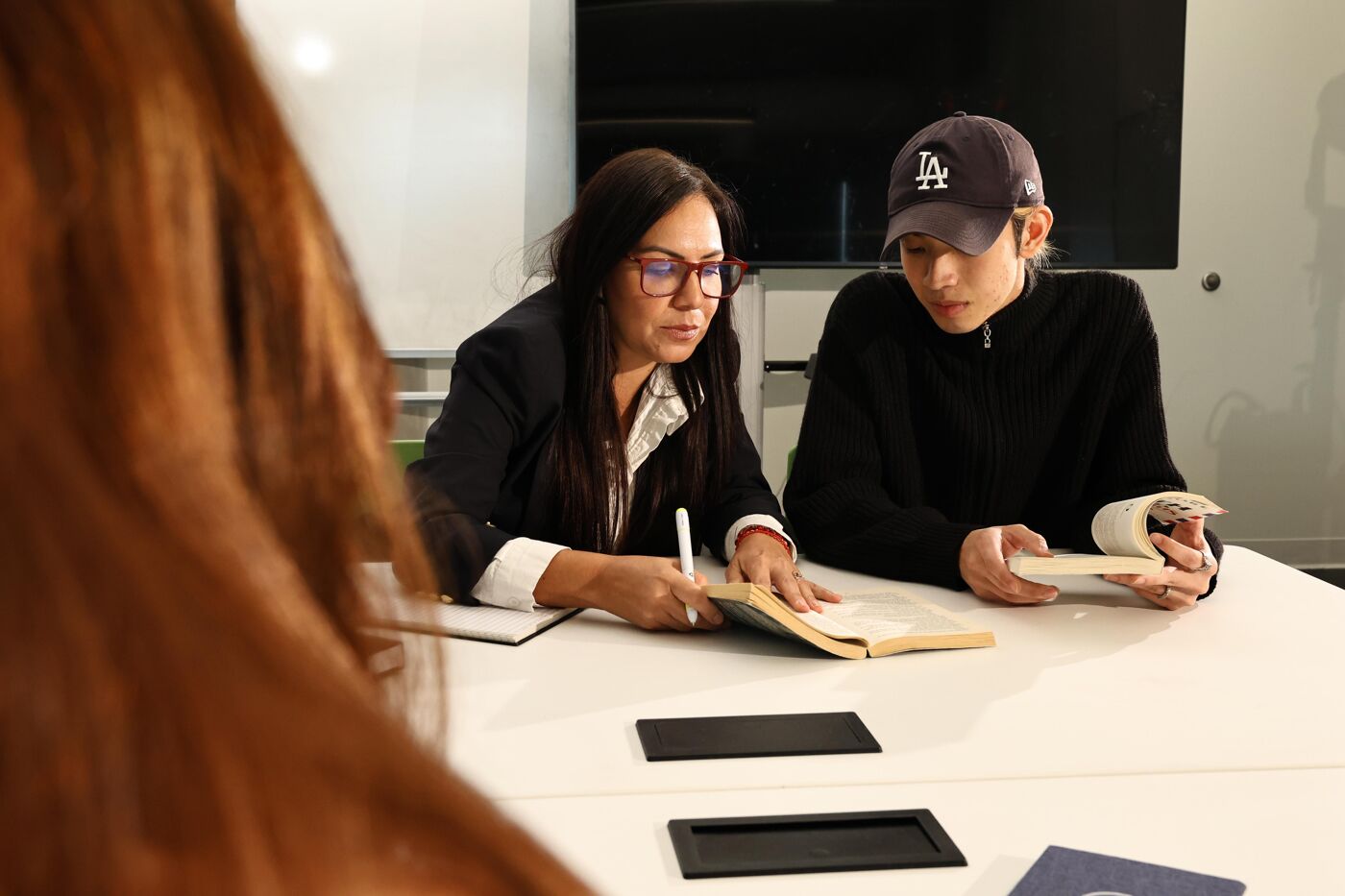 The image shows two people sitting at a table, seemingly engaged in a discussion or study session. One person is wearing a baseball cap and the other is wearing glasses. They both appear to be looking at a book or notebook on the table.