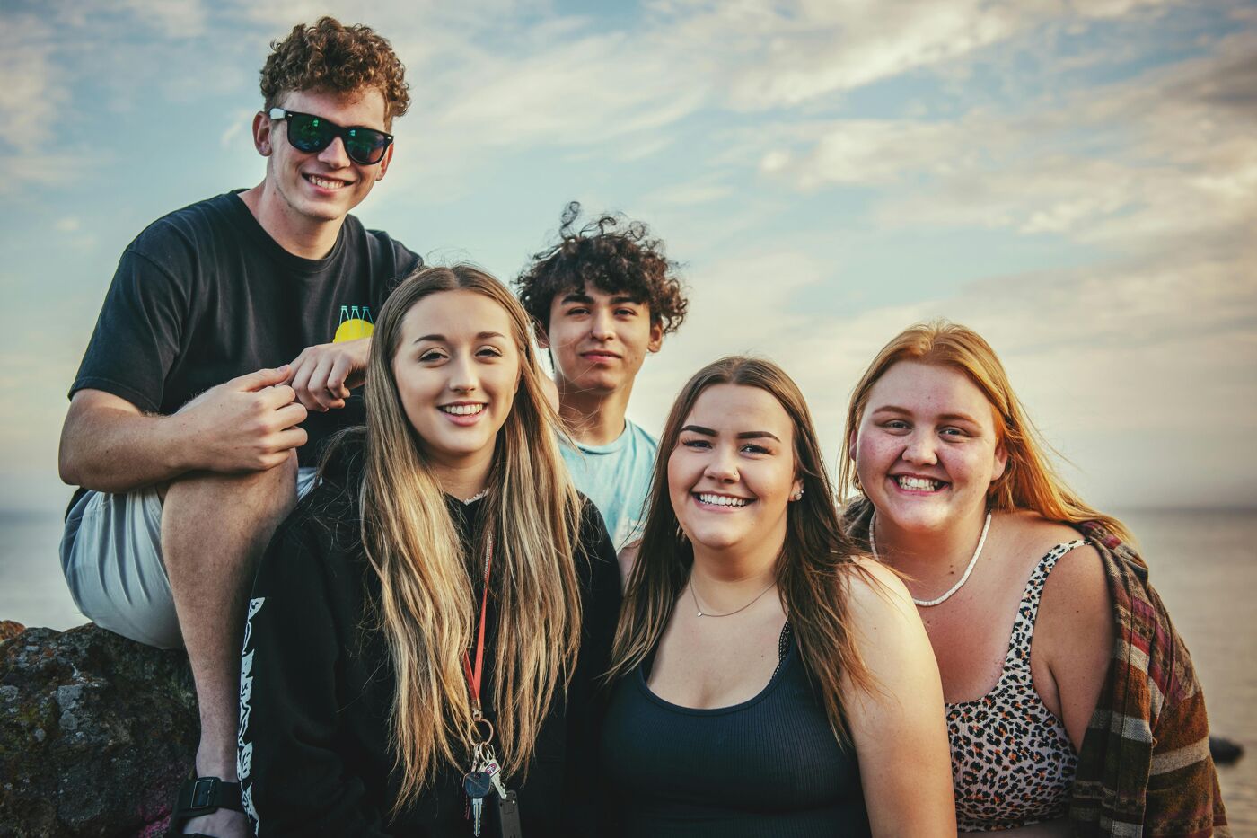 The image shows a group of five young people posing for a photo outdoors. They are smiling and appear to be enjoying themselves. The background includes a body of water and a cloudy sky.