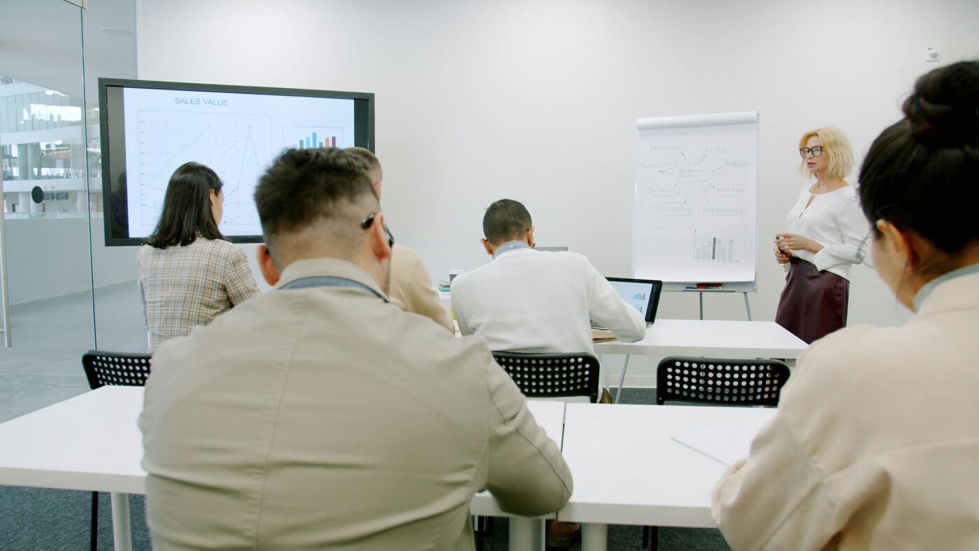 The image shows a business presentation in a conference room. A woman is presenting to a group of people who are seated at tables. A screen displays a graph, and a whiteboard is also present.