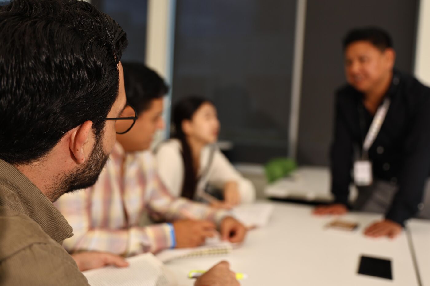 The image shows a group of people gathered around a table, seemingly in a meeting or discussion. One person is standing and addressing the others, who are seated and listening attentively. The setting appears to be an office or conference room.